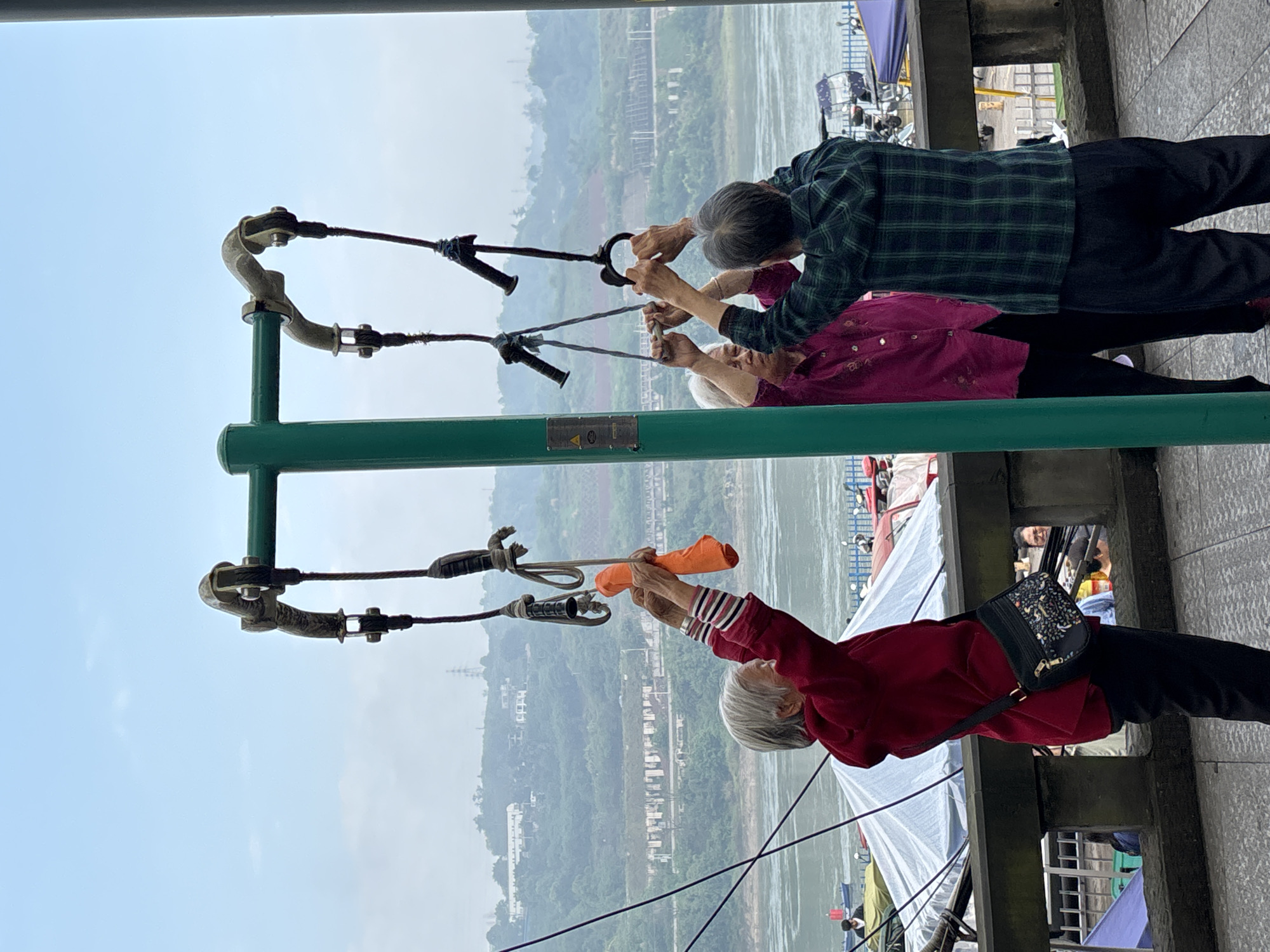 Three individuals are exercising outdoors in Jiangjin, China. They are utilizing a green metal frame with suspended hand grips, pulling on the grips as a form of exercise. The background reveals a river and surrounding hills. The weather appears overcast or foggy. The foreground shows a paved area where the individuals are standing.