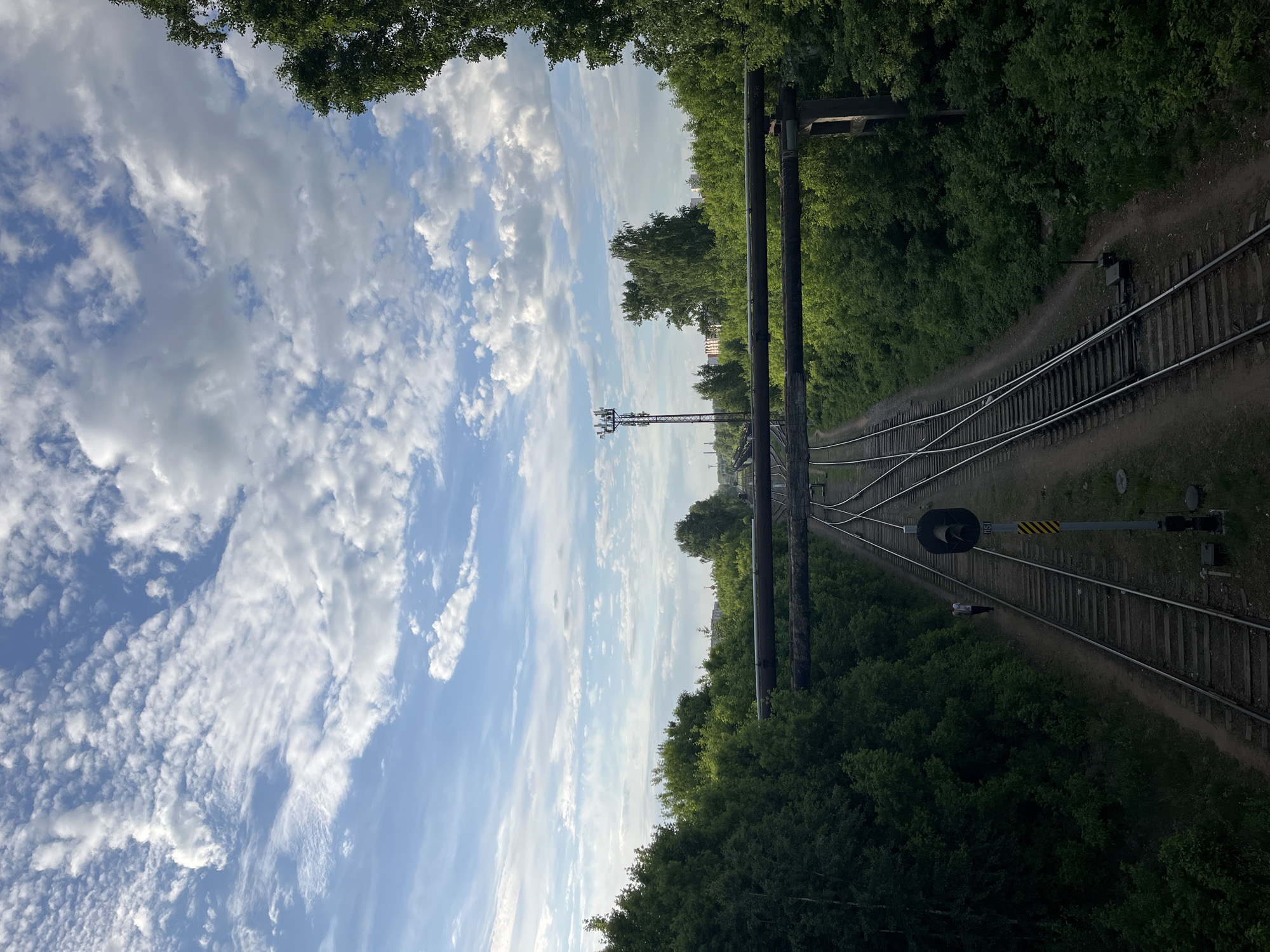 The image presents a view of a railway track in Chaikovsky, Russia. The track is set amid lush greenery, with trees lining both sides. Above, a sky filled with clouds creates a dynamic backdrop. A lattice tower and industrial structures are visible in the distance. The railway track has switches and signals. Above the railway track are some pipes.