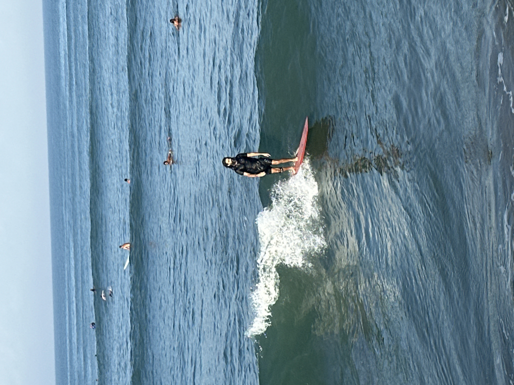 In North Kuta, Indonesia, a surfer is riding a wave near other surfers in the ocean. The surfer in the foreground is a male with dark, curly hair, wearing a black wetsuit and black shorts. He is standing on a red surfboard and is actively surfing on a breaking wave. The water is blue with white foam from the wave. In the background, other surfers are visible in the ocean, some paddling and some waiting for waves. The sky is clear and light blue.