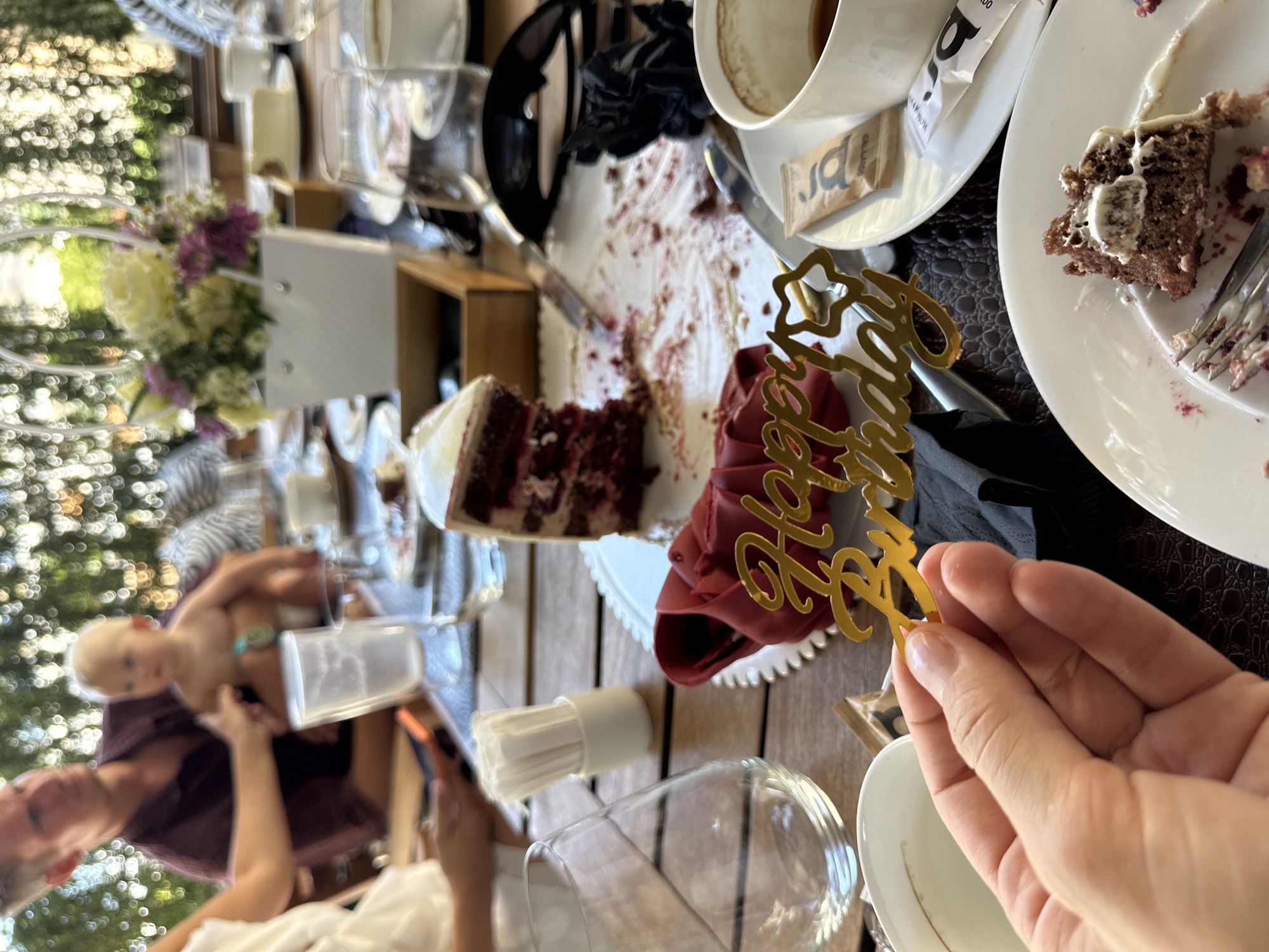 The image captures a table setting at a restaurant, likely outdoors, with a focus on remnants of a birthday celebration. In the foreground, a hand is holding a gold "Happy Birthday" cake topper. Behind it, on a white plate, is a slice of red velvet cake with white frosting, and scattered crumbs and red sauce. A red napkin is artfully folded to the side. To the right, a white teacup with a dark liquid is partially visible, next to a plate with a small packet of sugar. Further back, a white bag with a floral arrangement of purple and yellow flowers sits on the table. A partially eaten cake is also visible on another plate. In the background, a man wearing a maroon shirt is holding a baby. The table is made of wood. The overall atmosphere suggests a completed or near-completed birthday meal. The provided context indicates the location is Tivat, Montenegro.