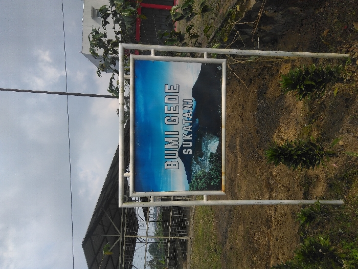 A rectangular sign with a white metal frame stands upright on dark brown, tilled earth. The sign displays an image of a dark mountain range with a lighter, possibly volcanic, feature on its lower slopes, and green vegetation in the foreground. White capital text on the sign reads "BUMI GEDE" and below it "SUKATANI." Three small, leafy green plants are rooted in the earth directly in front of the sign. To the left, a large, open-sided structure with a dark, sloped roof and metal truss framework is visible. Beyond this, a tiered structure or fence is partially seen. To the right, a white multi-story building with a red base is partially obscured by a tree with dark green foliage. A tall, thin, dark pole extends vertically from the ground into the sky, slightly to the right of the sign. A thin, dark horizontal wire crosses the upper sky. The sky is overcast with gray and white clouds. The scene is located in Sukatani, Indonesia.