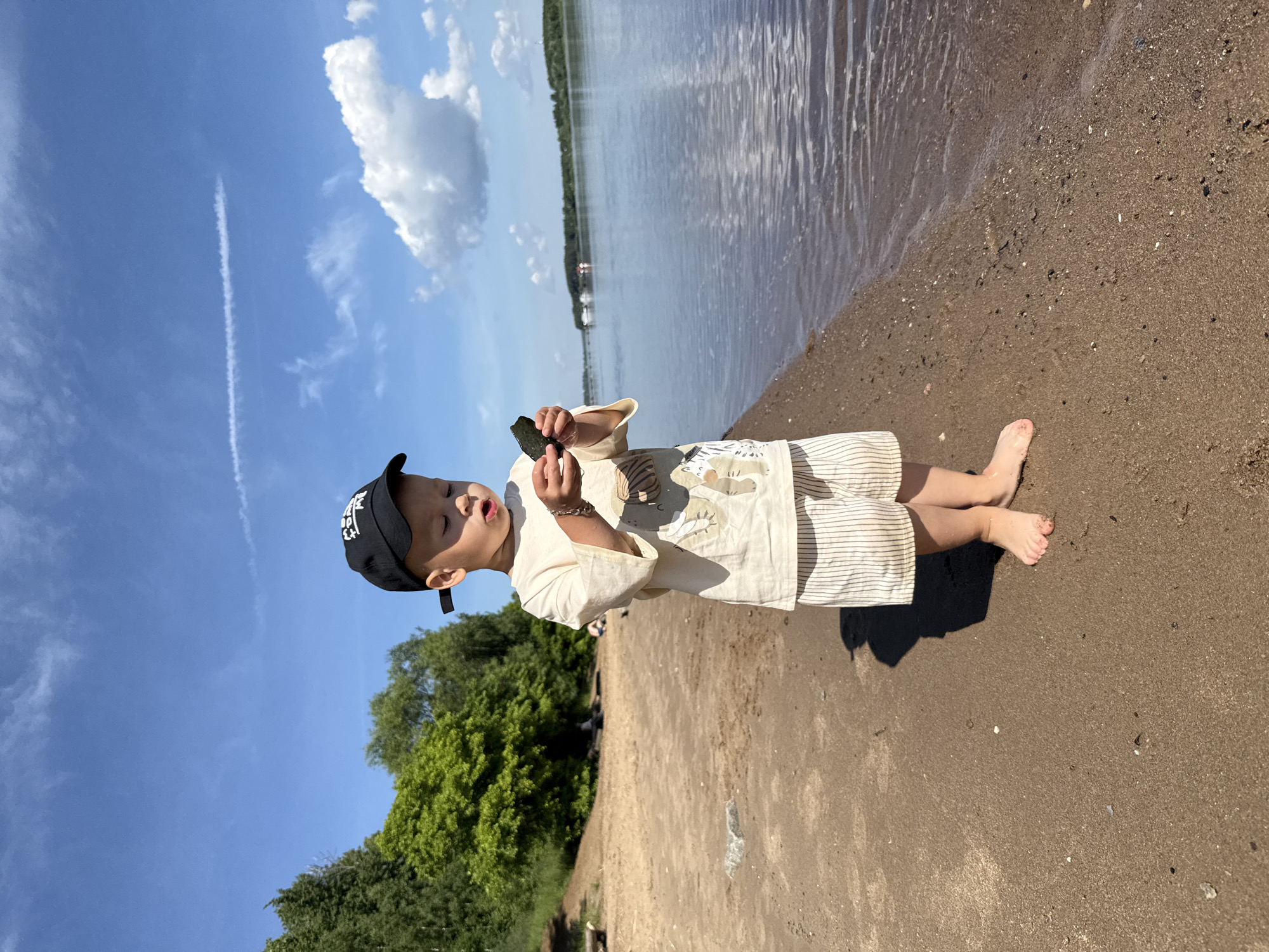 A young boy wearing a black baseball cap, a light-colored t-shirt with a graphic print, and striped shorts stands barefoot on a sandy beach. He is holding a dark-colored object in his hands and looking down at it. The boy is positioned near the edge of a body of water, with ripples visible on the surface. In the background, there is lush green foliage and trees along the shoreline, with a bright blue sky dotted with white clouds above. The scene appears to be outdoors in a natural setting, possibly a park or a riverside.