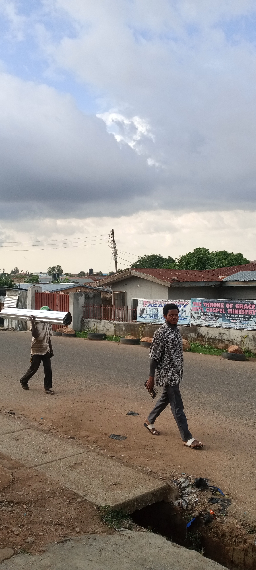 The image shows two men walking on a road in Jos, Nigeria. One man carries several metal pipes on his shoulder along the sidewalk. The other man walks on the road carrying a small, dark object, possibly a wallet or phone, in his hand. Behind them is a low building with signs indicating "PRECIOUS STON..." and "THE THRONE OF GRACE... GOSPEL MINISTRY". The sky is overcast with dark clouds. A storm drain is visible in the lower foreground, filled with trash. The environment appears to be a residential or mixed-use area in a city.