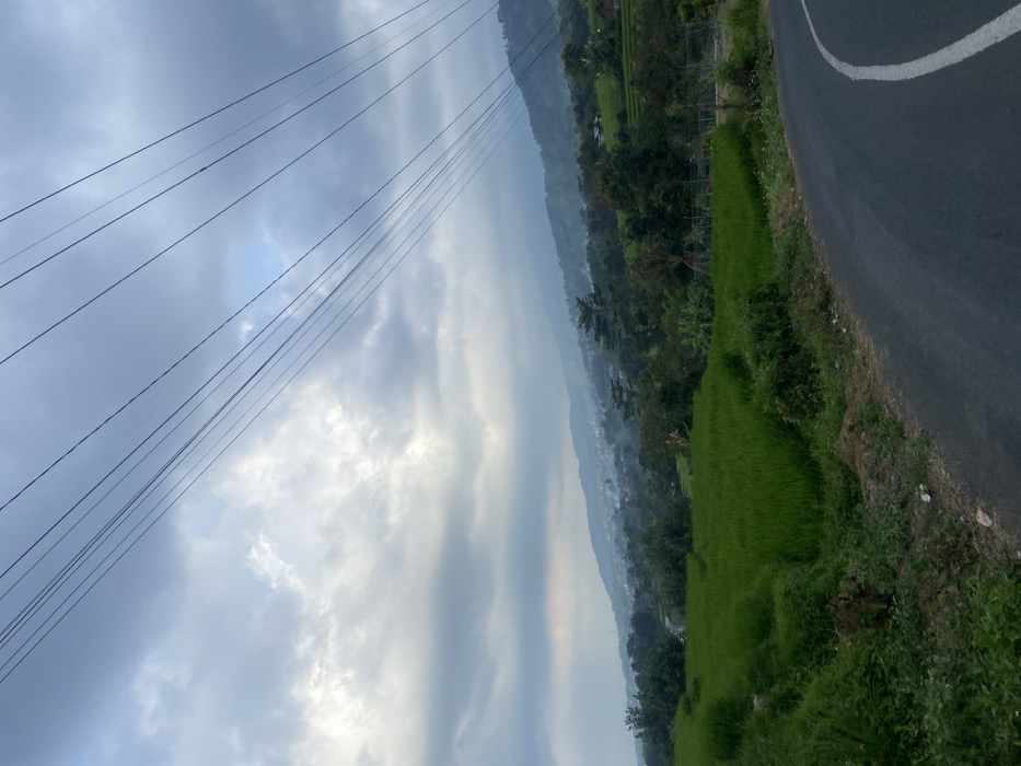 The image displays a static landscape scene in Sukoreno, Indonesia, under an overcast sky. An asphalt road, dark grey with a solid white line along its right edge, curves from the lower right corner towards the center of the frame. To the left of the road and extending into the mid-ground are vibrant green agricultural fields, indicative of rice paddies. Beyond these fields, the terrain ascends into hills and mountains, featuring scattered residential structures and dense foliage. A light atmospheric haze or mist is present over the distant hills and inhabited areas. Multiple dark, thin power lines traverse diagonally across the upper portion of the sky, from the upper left to the upper right. The sky is dominated by varied shades of grey and white clouds. No human figures, specific actions, or interactions are visible within the frame.