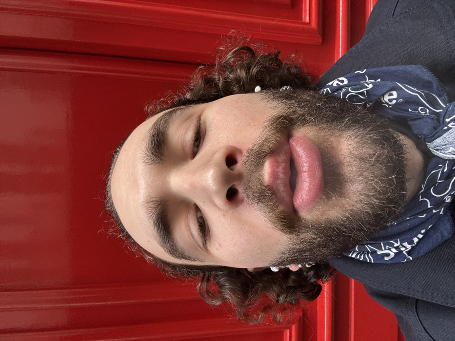 The image displays a close-up portrait of a person, rotated 90 degrees clockwise. The subject's head is tilted to the right, with their face occupying the central and lower-left portions of the frame. The background is a vibrant, solid red surface, possibly a paneled door or wall, which extends across the top and left sides of the image.
The person has curly dark brown hair, which is visible around the sides of their head and forehead. They have a thick, dark beard and mustache covering their chin, cheeks, and upper lip. Their skin appears light-toned. Their eyes are partly closed or looking downwards, with visible eyelids and well-defined dark eyebrows. A small dark piercing is visible in the left nostril. The lips are full, slightly parted, and show a pinkish-red hue with visible texture.
Small, round, white or silver bead-like earrings are visible on both ears, nestled within the curly hair. Around the person's neck, on the right side of the frame, a dark blue bandana or scarf with a white, intricate pattern (resembling paisley or a similar design) is tied. Part of a dark-colored garment, possibly a jacket or shirt, is visible on the far right, underneath the bandana.
The overall expression is relaxed or contemplative due to the partly closed eyes and slightly parted lips. No discernible activity is taking place other than posing for the photograph. There is no clearly legible text visible in the image, though the pattern on the bandana is intricate.
