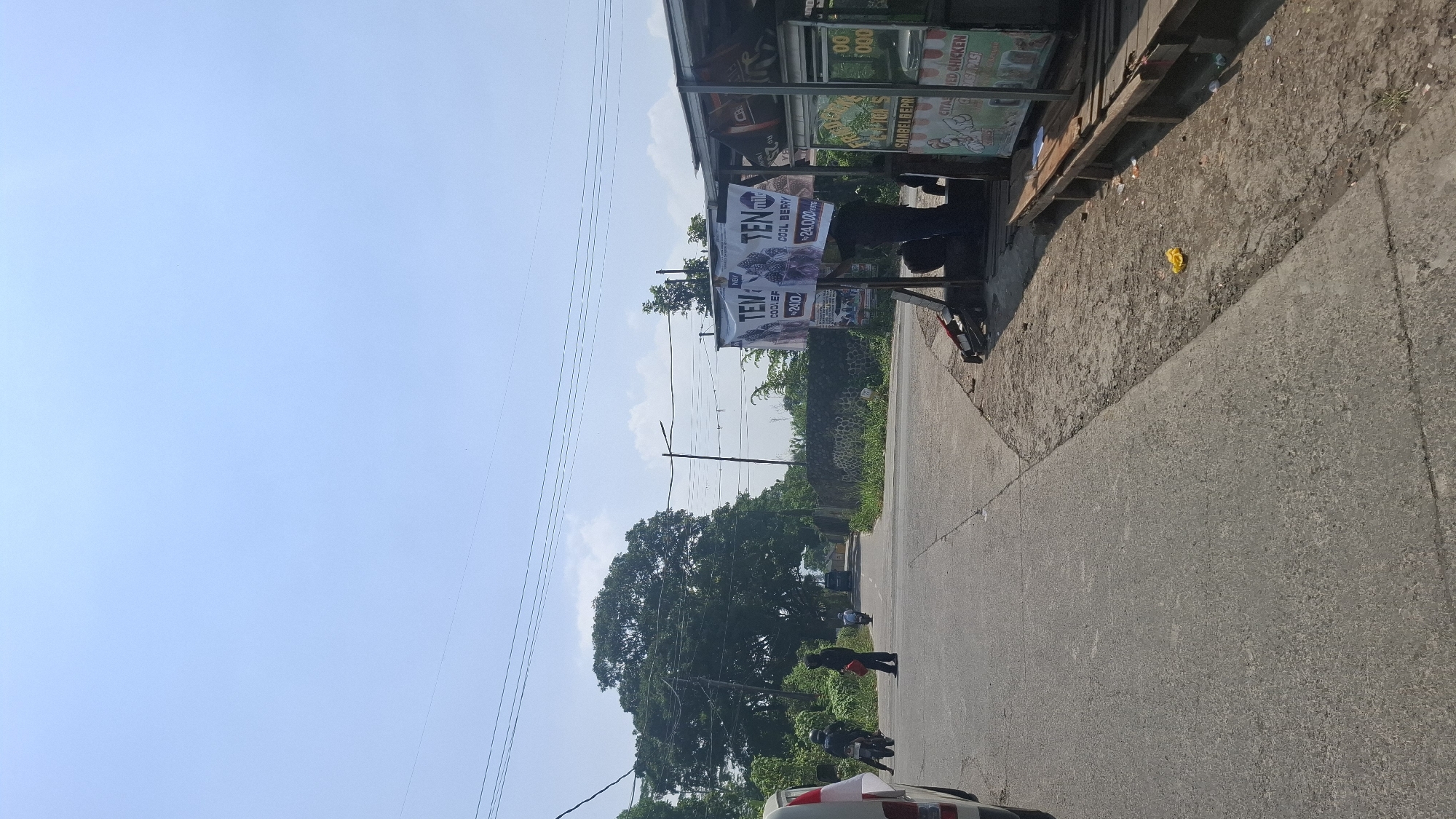 The image shows a roadside scene in Solokpandan, Indonesia. The foreground features an asphalt road with a slight incline. To the right, there is a small food stall with a corrugated metal roof and wooden supports. The stall displays various items, including signs that read "FRIED CHICKEN" and "00 FOOD". A person wearing dark clothing is standing behind the stall. Two banners are attached to a pole in front of the stall, one advertising "TEN COOLER BERRY" for "Rp 24.000".
In the middle ground, the road continues, bordered by overgrown vegetation and a stone wall on the right. Several individuals are visible on the road. Two people on motorcycles are proceeding away from the viewer, and another person is standing on the side of the road holding a red bag. A large, leafy tree dominates the left side of the middle ground.
The background consists of a bright, clear sky with a few scattered clouds and power lines stretching across the frame. The overall environment suggests a rural or semi-urban setting.