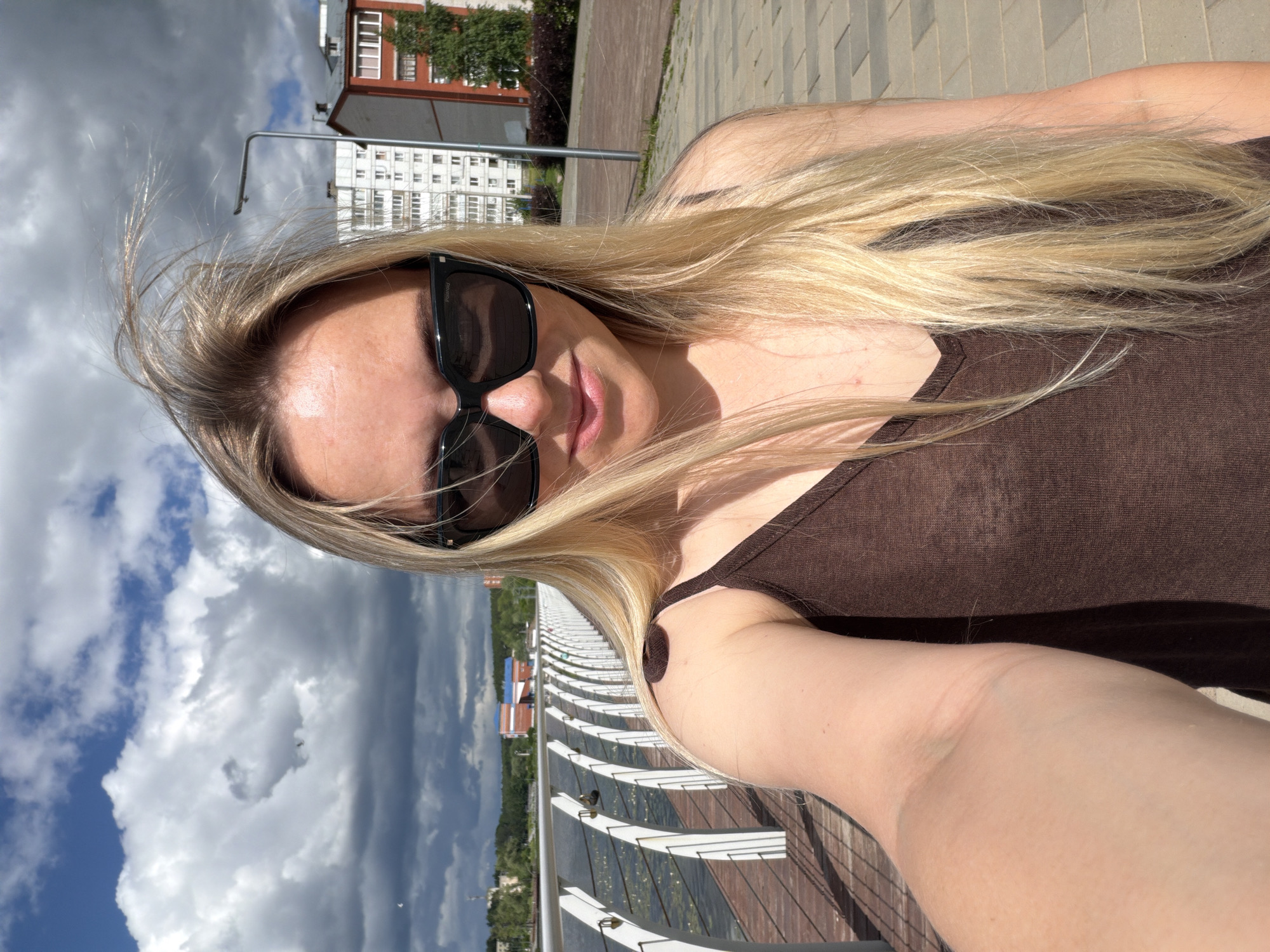 A young Caucasian woman with blonde hair and sunglasses is taking a selfie outdoors. She is wearing a brown tank top and is standing on a paved walkway next to a body of water. In the background, there is a railing with white vertical supports, buildings, trees, and a cloudy sky. The location is Votkinsk, Russia, specifically on the embankment of the Kama River.