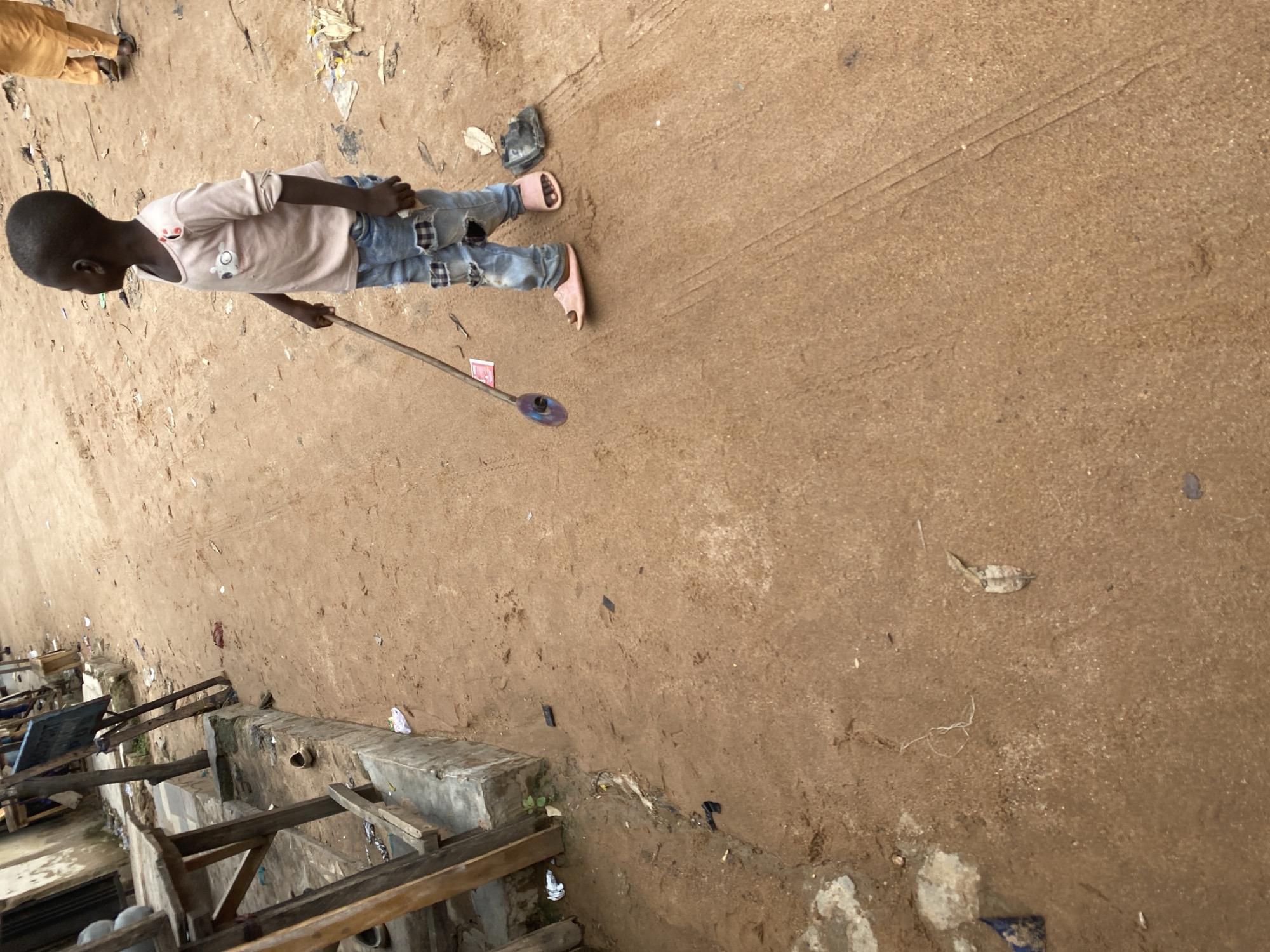 A young Black child, wearing a pale pink t-shirt with a cartoon animal graphic and distressed blue jeans, stands on a dirt path. The child is holding a long, thin stick with a round, purple object attached to the end. The child is facing left in the frame. Nearby, another person, wearing tan pants, is partially visible at the top left of the image. The ground is covered in a light brown dirt, with some scattered debris such as leaves and small pieces of trash. Tire tracks are visible in the dirt. In the background, to the left, are the partial structures of what appear to be market stalls or shelters made of wood and concrete. The scene appears to be outdoors. The lighting suggests it is daytime.