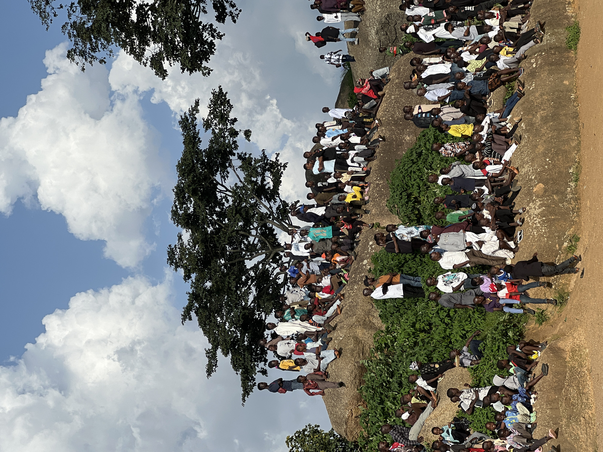 In the city of Jos, Nigeria, a large group of people are gathered on and around a large rock formation. The individuals are of various ages and genders, dressed in casual clothing. Some are sitting on the rock, while others are standing or leaning against it. A large tree with dark green foliage is situated behind the rock formation. The sky above is partly cloudy with fluffy white clouds against a clear blue backdrop. The environment appears to be an outdoor, natural setting, possibly a scenic overlook or a gathering point. The overall scene suggests a community event or a casual get-together, with people enjoying the elevated vantage point.