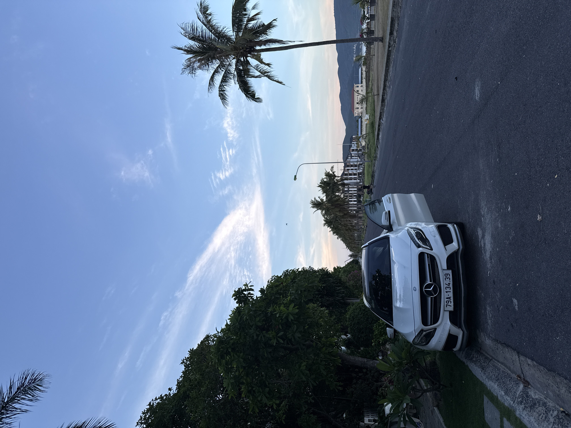 A white Mercedes-Benz GLA-Class parked on the side of an asphalt road in a coastal city. The car is facing forward with its driver's side door open. The license plate reads "79A-134.39". To the left of the car is a patch of green grass with some plants. The road stretches into the distance, lined with palm trees and buildings. In the background, there are mountains and a body of water. The sky is blue with some wispy clouds, and the sun appears to be setting. The scene is peaceful and serene.
The location is likely in the city of Nam Nha Trang Ward, Vietnam, given the presence of palm trees, coastal scenery, and the Vietnamese license plate format. The overall atmosphere suggests a leisurely evening stroll or a stop by the beach.