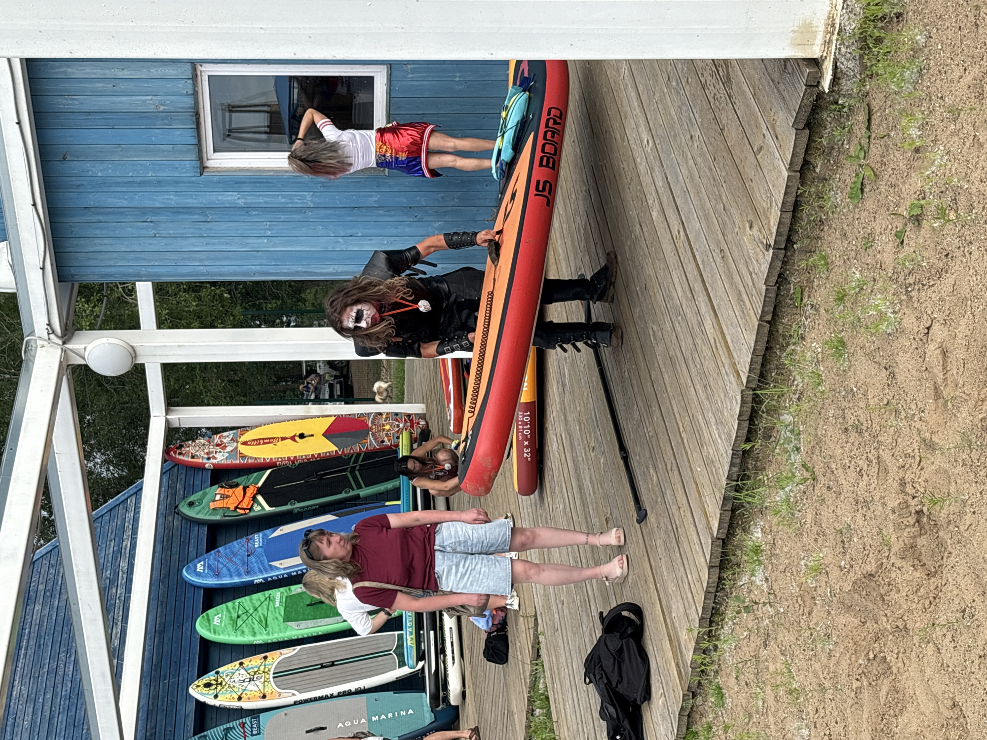 A group of people are gathered on a wooden deck outside a blue building with a white roof structure. In the background, a row of colorful paddleboards are propped against the building. On the deck, a woman in a burgundy t-shirt and denim shorts stands, looking to the side. To her right, a person dressed in a black, leather-like costume with white face paint, resembling a character from "The Joker," holds a red paddleboard. Another woman with gray-streaked hair, wearing a white shirt and a colorful skirt, stands in a doorway of the blue building, touching her hair. The environment appears to be outdoors, with sand and some vegetation on the ground in the foreground. The scene suggests a recreational area, possibly a beach or a watersports center.