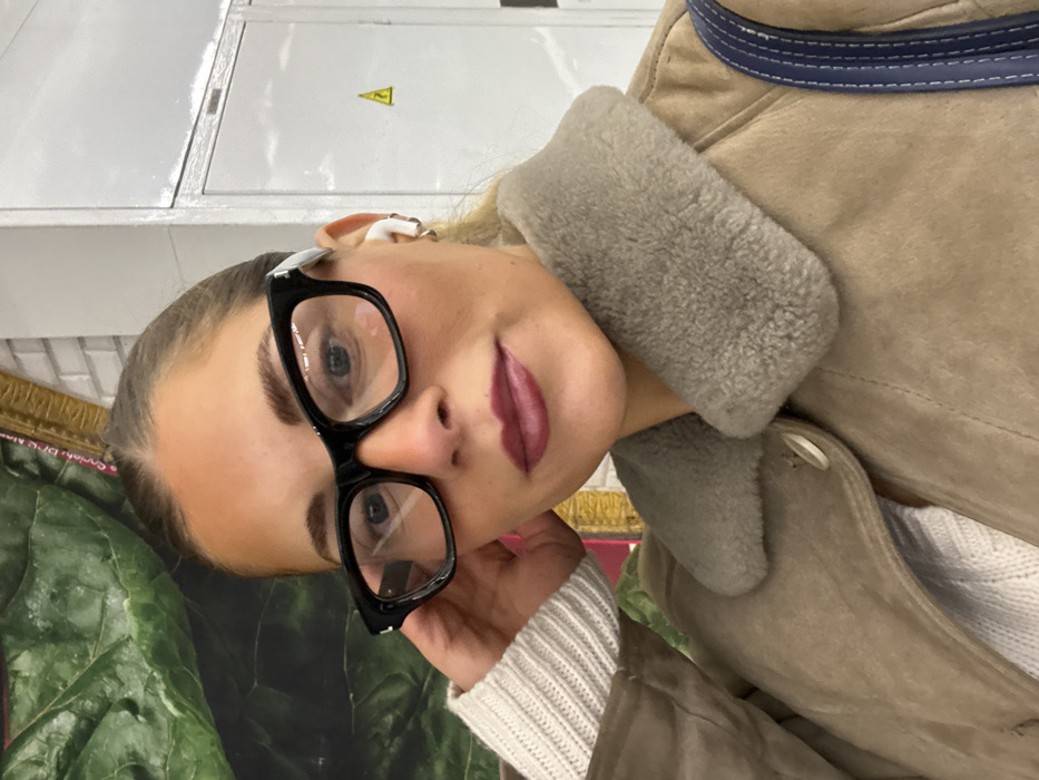 A close-up, slightly tilted portrait shot captures a young woman from the chest up. She is looking directly towards the viewer with a subtle smile on her face. Her light-colored hair is pulled back, revealing a white earbud in her right ear. She wears stylish black-rimmed glasses, through which her light-colored eyes are visible. Her eyebrows are well-defined, and her lips are colored with a matte, dark berry or plum lipstick.
She is dressed in a light brown or tan jacket, possibly suede or shearling, featuring a thick, fluffy light grey/beige collar. A distinctive detail on the jacket is a dark blue strap or trim with white stitching, visible on her upper right shoulder area. Beneath the jacket, a white, ribbed knit sweater is visible on her right arm. Her left hand is subtly raised, with her fingertips gently touching her jawline near her ear.
The background is somewhat indistinct but offers a few details. To her upper left (our right), there's a light-colored, possibly white, surface that resembles a wall or a large panel, with a small yellow triangular sign with an exclamation mark icon on it. To her lower left (our left), a large, vibrant green, leafy pattern is visible, which could be part of a poster, artwork, or fabric. Adjacent to the green pattern, there's a textured gold or bronze element. The overall lighting appears soft and even.