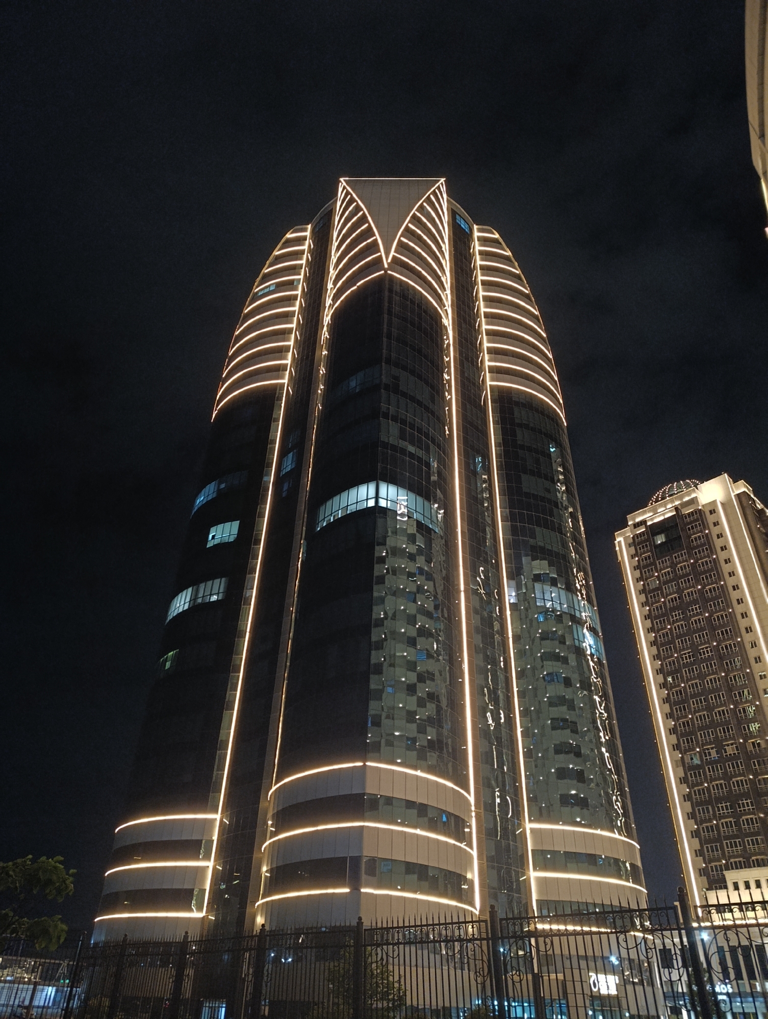 The image shows a night view of the modern buildings in Grozny, Russia. In the foreground, a tall, curved skyscraper with glass facade and illuminated horizontal bands of light is prominently featured. The skyscraper has a unique design with vertical elements that accentuate its height. To the right of this main building, another taller, more rectangular skyscraper with a similar illuminated facade is visible. The sky is dark, indicating nighttime. A black metal fence runs across the bottom of the image. The lighting on the buildings suggests they are illuminated for aesthetic purposes at night.