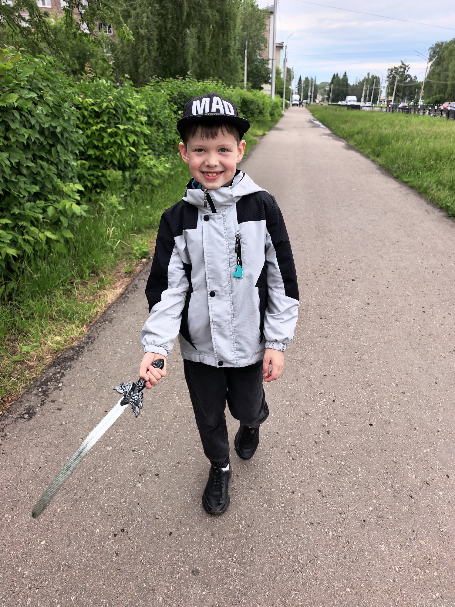 A young boy, appearing to be around 6-8 years old, is captured walking on a paved path, smiling broadly at the camera. He has short, dark hair and a joyful expression, revealing his teeth.
He is dressed in a casual outfit consisting of a black baseball cap with the word "MAD" prominently displayed in white letters on the front. He wears a grey and black color-block hooded jacket, black pants, and black sneakers. A small, light blue dinosaur-shaped keychain hangs from a zipper on his jacket. In his right hand, he holds a detailed toy sword with a silver blade and an ornate, dark hilt, pointing it downwards and slightly forward.
The scene takes place outdoors on a paved pathway. To the boy's left, a dense wall of vibrant green bushes and trees lines the path, with a building partially visible through the foliage in the distance. To his right, a strip of green grass separates the path from a street, where utility poles, distant vehicles, and more trees can be seen under an overcast sky. The setting appears to be an urban or suburban street or park pathway in Sterlitamak, Russia.
The boy's cheerful smile and the toy sword suggest a playful and happy moment, possibly during a walk or imaginative play. The time of day is daytime, under a cloudy sky.