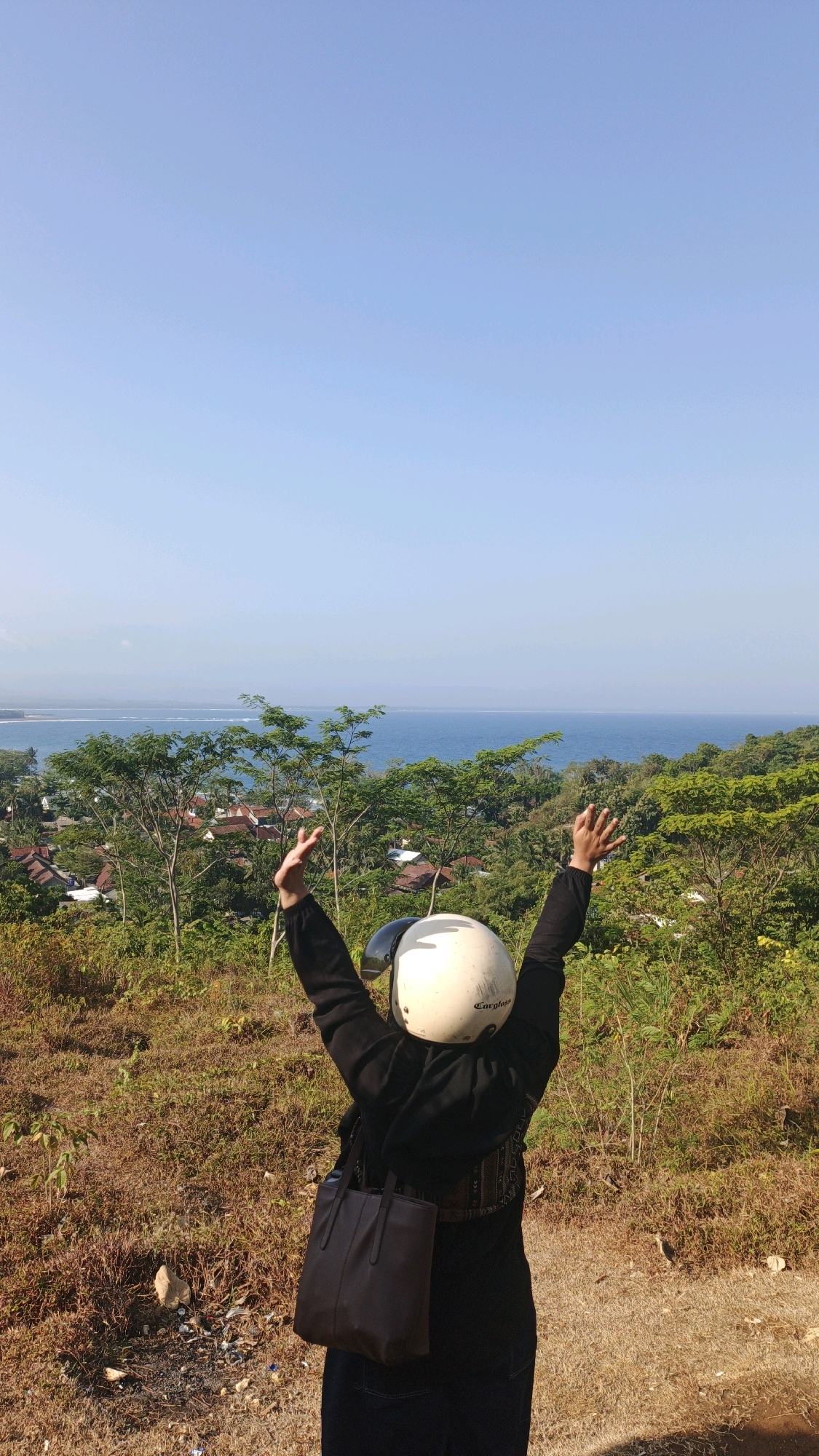 A person wearing a black hoodie and a white helmet stands on a dirt path, facing away from the camera. Their arms are raised in the air. The person is carrying a dark brown bag over their left shoulder. Behind them, a lush green landscape slopes down towards the blue ocean. In the distance, the ocean meets the horizon under a clear blue sky. Small buildings with red roofs are visible amongst the trees on the hillside. The ground in the foreground is covered in dry grass and sparse vegetation. The overall scene depicts a scenic overlook with a person expressing joy or freedom. The location is Desa Batukaras, Indonesia.