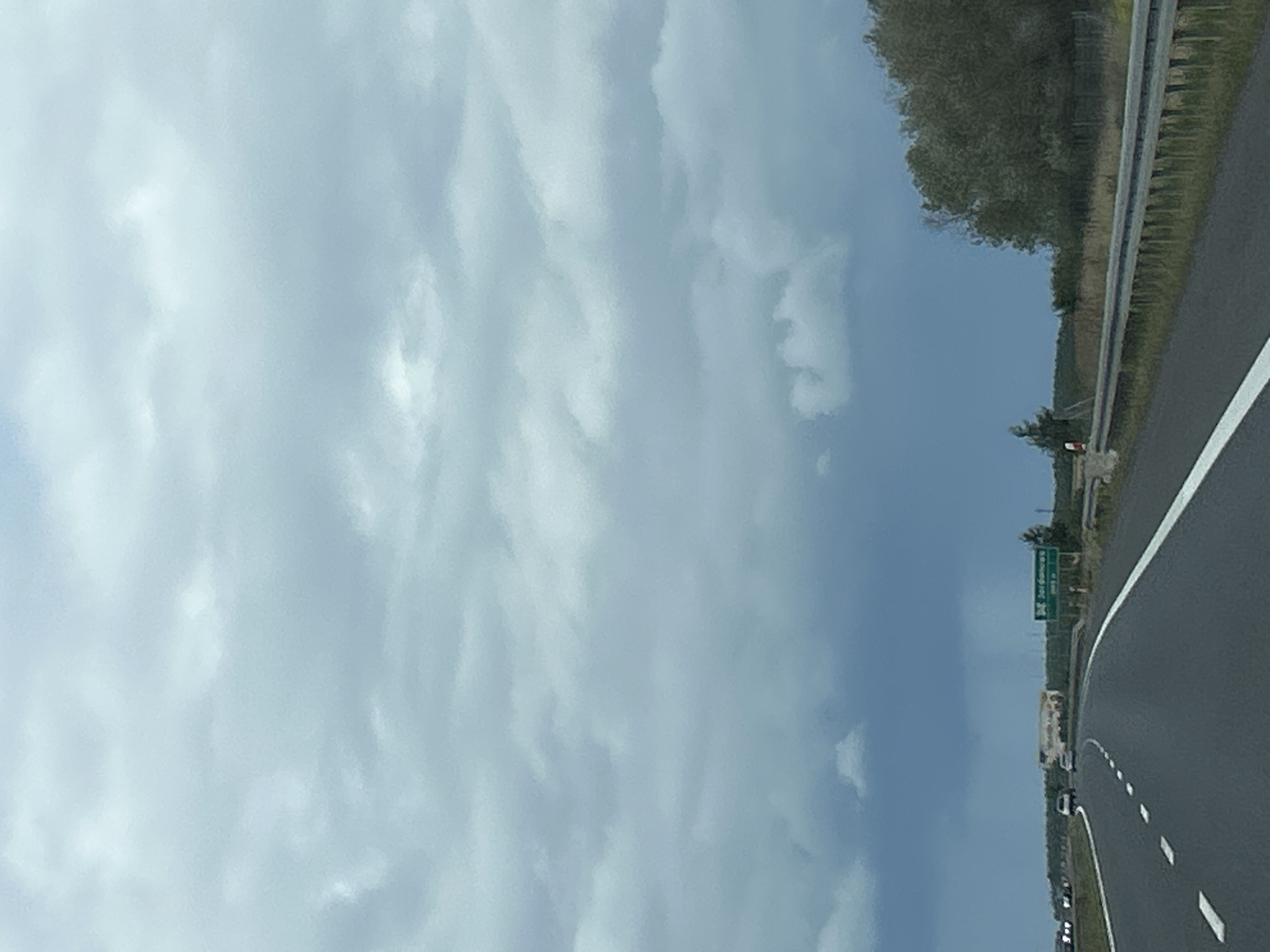 The image shows a highway scene on a cloudy day. A road stretches into the distance, with white lane markings and a metal guardrail on the right. Several cars are visible on the highway, driving in the same direction. On the right side of the road, there are trees and vegetation. A green highway sign is visible in the distance on the right, indicating "Jordanowo" and "500 m". The sky is overcast with dramatic, dark clouds suggesting impending rain. The overall impression is one of travel on a highway under a stormy sky.