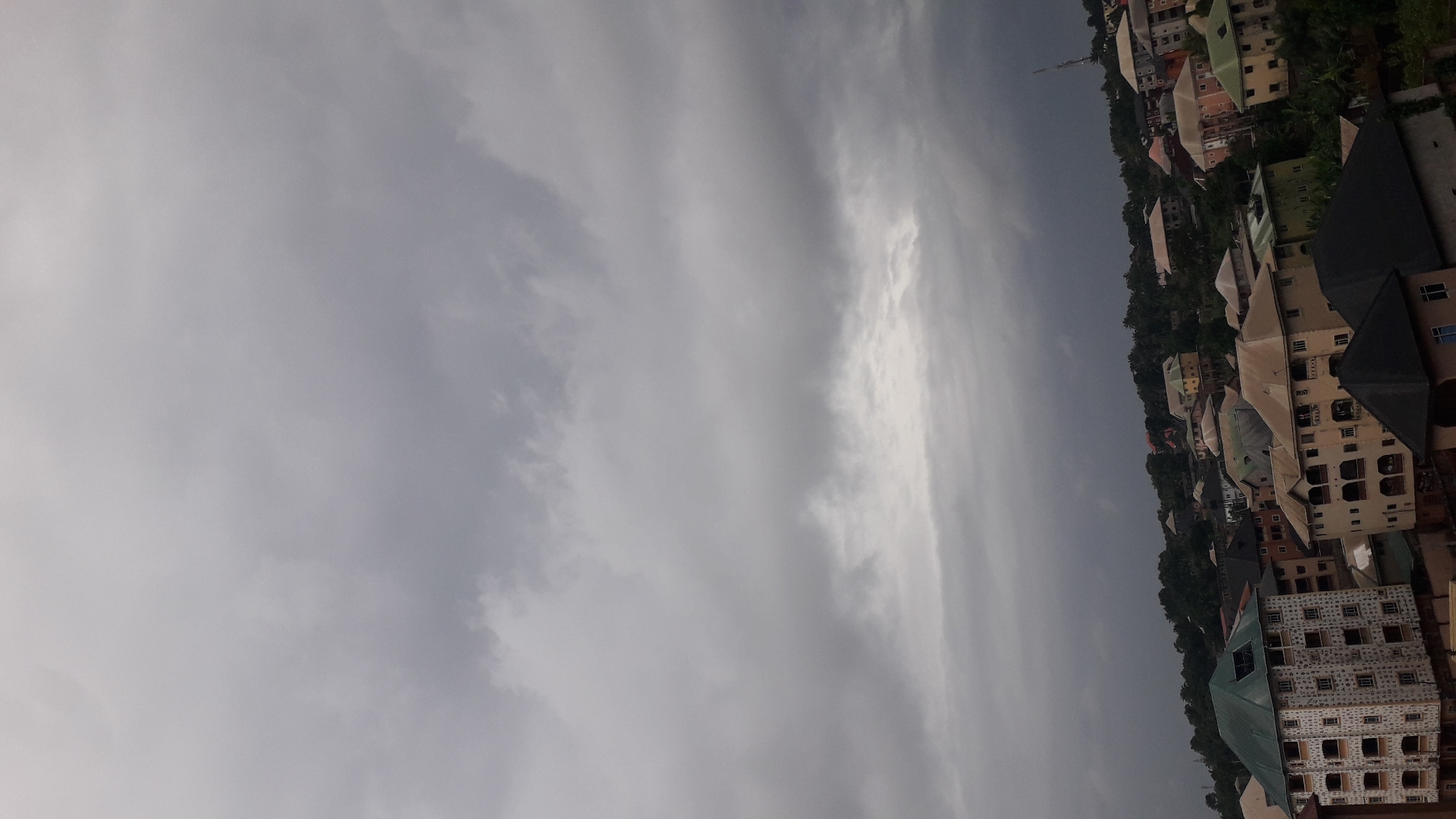 A high-angle, full shot captures a cloudy, overcast sky above a densely packed residential area in Nkpor, Nigeria. The sky is dominated by thick, gray clouds, suggesting a dull or stormy day. Below the clouds, a sprawling cityscape unfolds. Numerous multi-story buildings, with varying architectural styles and colors, are clustered together on a hillside. Some buildings feature prominent balconies and rooftop structures. Trees and vegetation are interspersed among the buildings, with denser foliage appearing on the higher parts of the hill. The overall impression is of a vibrant, lived-in urban environment under a brooding sky.