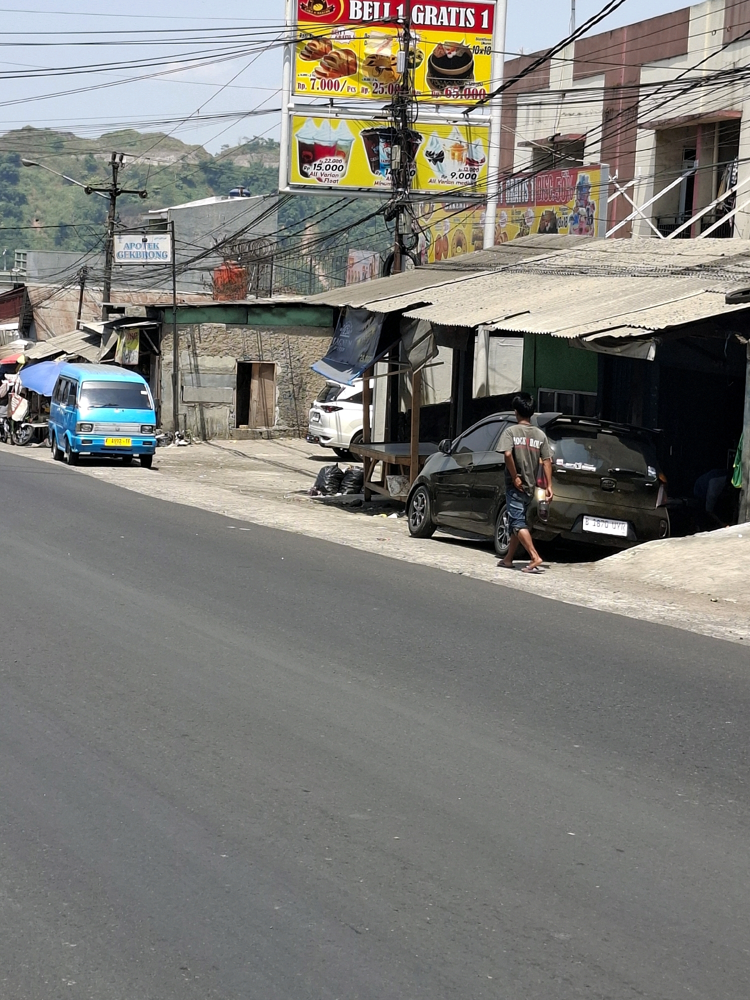 The image shows a street scene in Cikahuripan, Indonesia. In the foreground is a paved road. On the right side of the road, a dark colored hatchback car is parked facing away from the camera. A man wearing a t-shirt and denim shorts is walking away from the car towards the right side of the image. On the left side of the road, a blue mini-van is parked facing the camera. Further back on the left is a small building with a sign that reads "APOTEK GEKBRONG". Above the buildings, a cluster of electrical wires is visible against a hazy sky. In the background, green hills are visible. The scene appears to be a commercial area with various shops and storefronts lining the street.