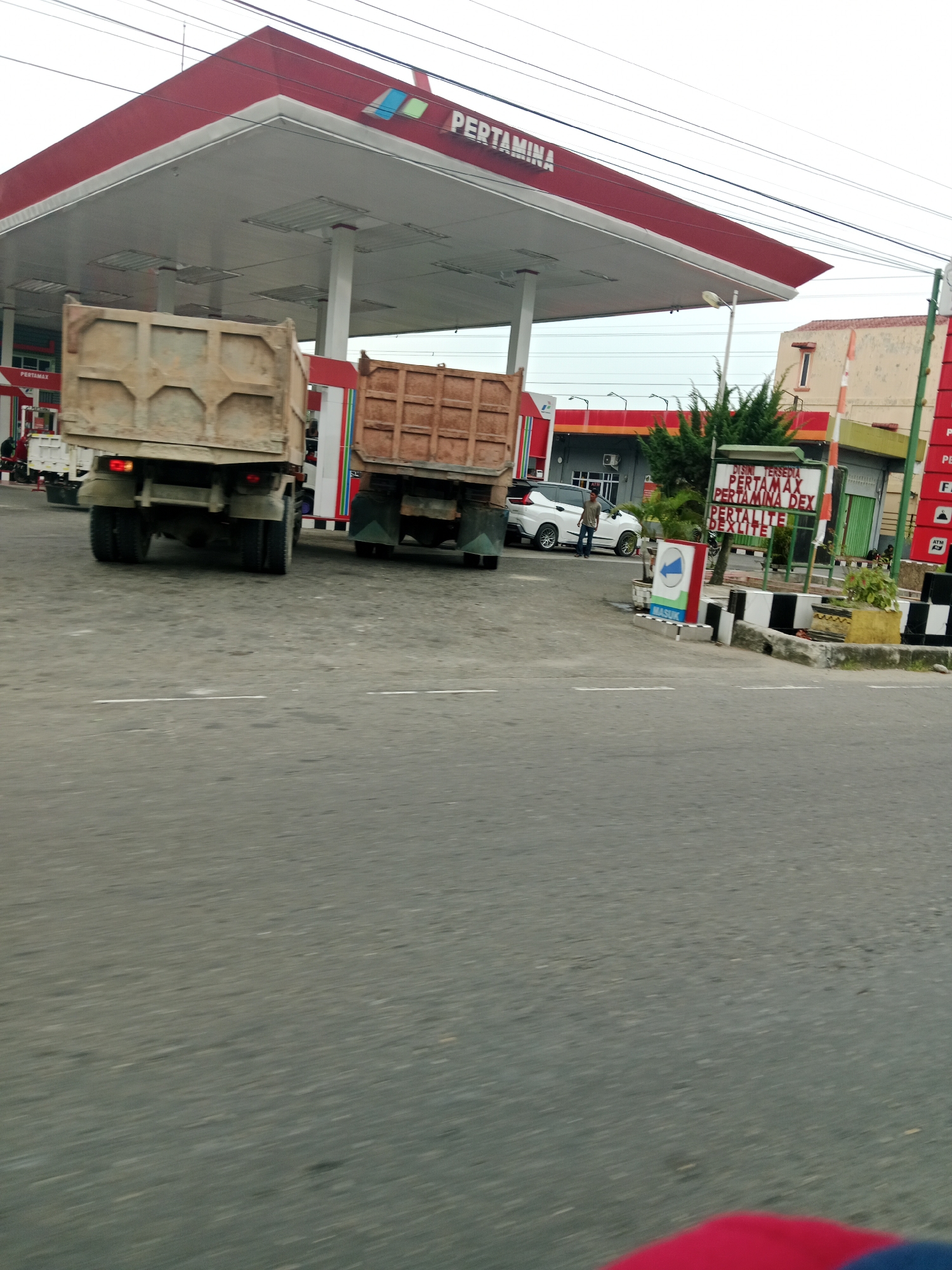 A daytime, slightly blurred exterior shot shows a Pertamina gas station in the city of Binjai, Indonesia. The gas station has a red roof with white trim. Two dump trucks are parked at the station, their beds raised. A white car and a few people are visible near the pumps. A sign lists available fuel types: Pertamax, Pertamina Dex, Pertalite, and Dexlite. The road in the foreground is asphalt with white lane markings. The sky is overcast.