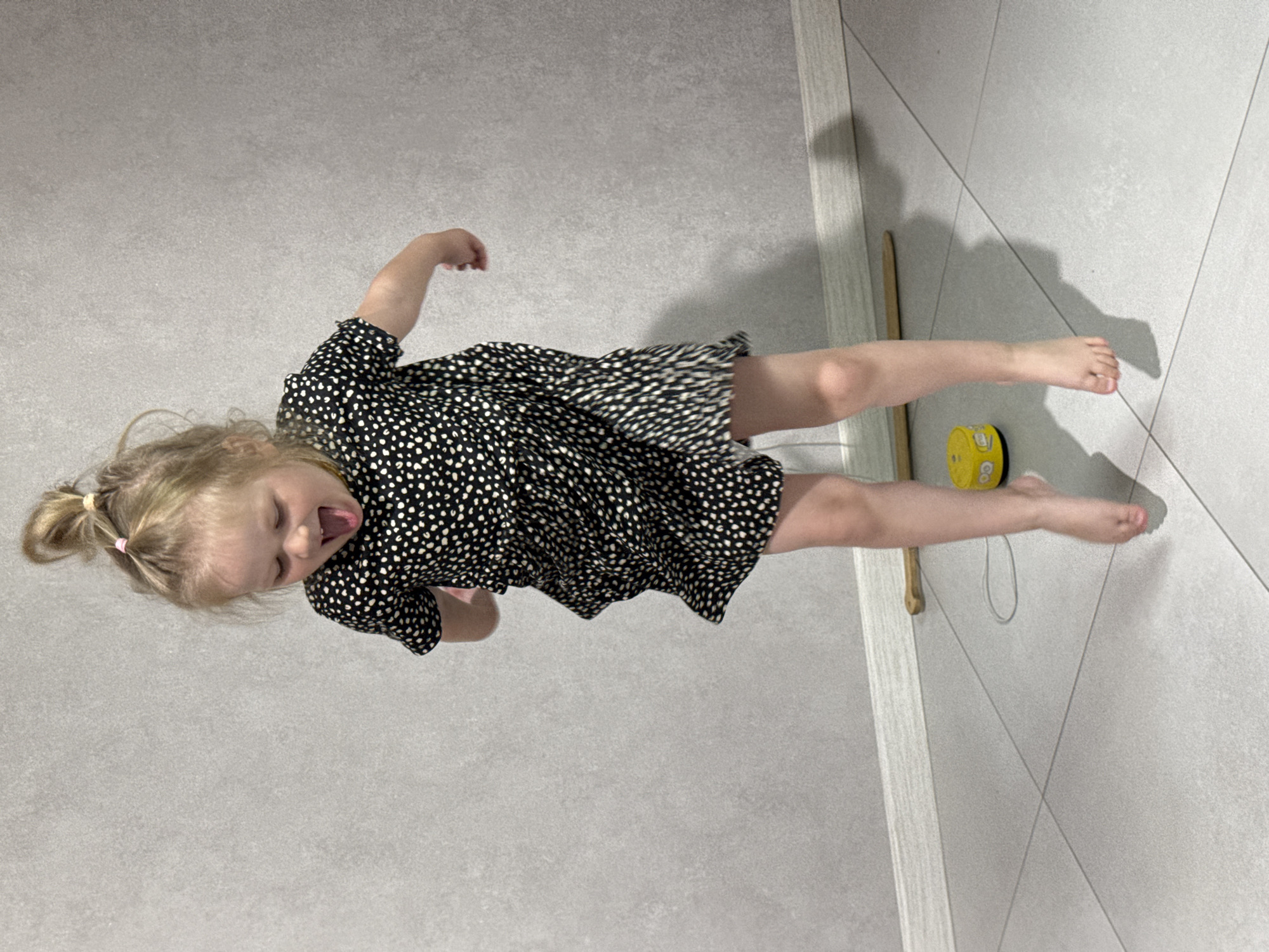 A young child with blonde hair pulled into a ponytail is lying on a tiled floor. The child is wearing a dark dress with white polka dots, and her mouth is open as if she is shouting or laughing. To the right of the child are a yellow toy drum, a drumstick, and a white cord. The location appears to be indoors, likely a residence in Gornyi (Dvurechenskoe S/P), Russia, as specified. The flooring is a light gray tile. The child's pose suggests play or movement.