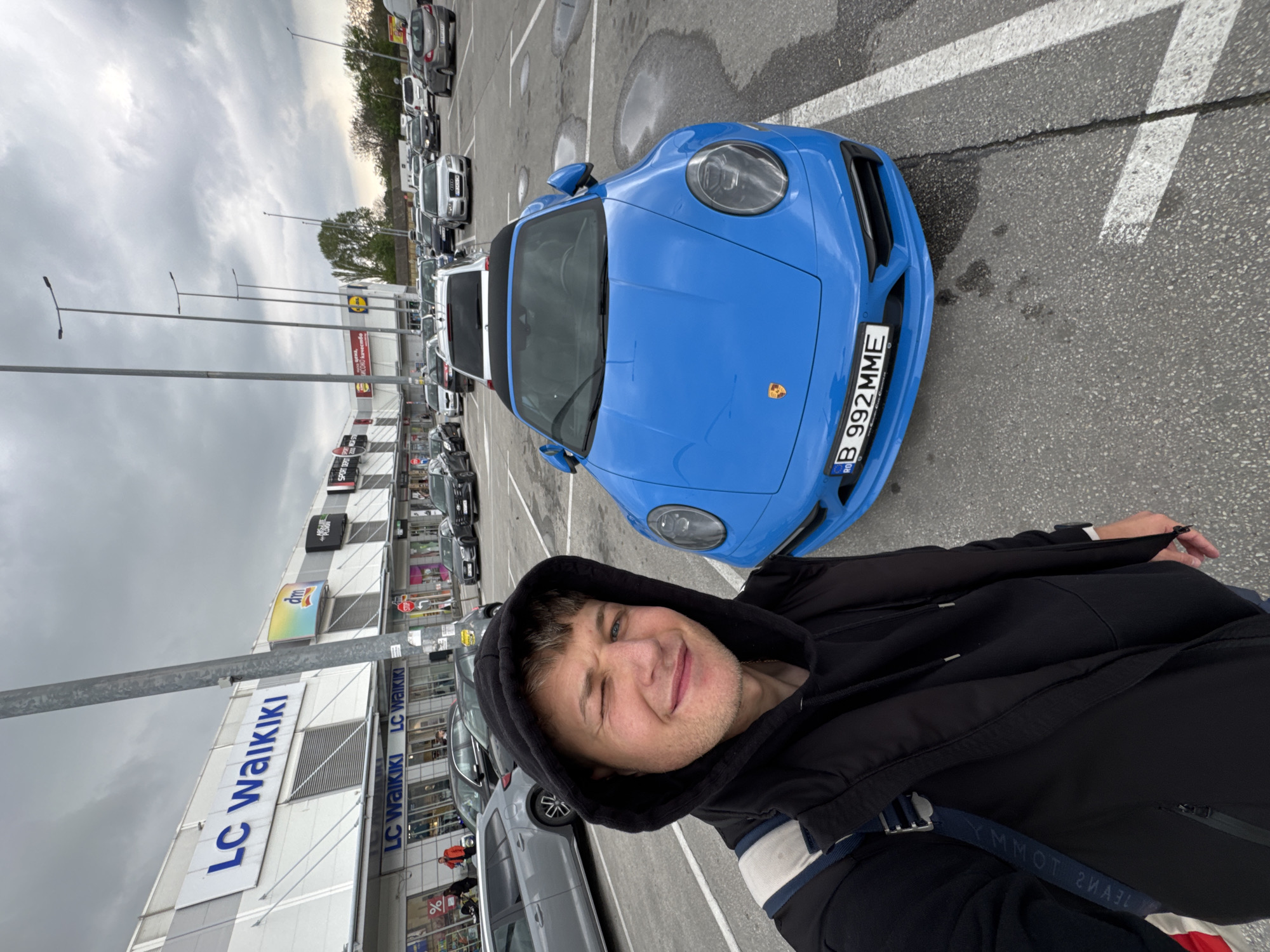 A person with a hooded jacket is taking a selfie in a parking lot, with a blue Porsche car in the background. The license plate on the Porsche reads "B 992 MME." Behind the car, there's a line of parked vehicles. In the background, signs for "LC Waikiki," "SPORT DEPOT," "LIDL" and other retail stores are visible. Based on the store names and the license plate on the car, the location is likely a shopping complex in Plovdiv, Bulgaria. The sky is overcast.