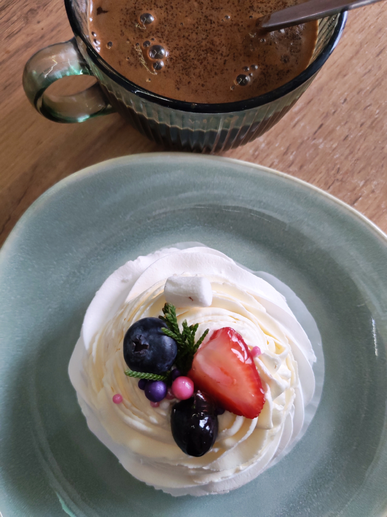A close-up shot captures a cup of coffee and a dessert. The coffee cup, made of green glass, sits at the upper part of the frame and contains coffee with bubbles on its surface. A spoon rests inside. Below, on a light blue plate, is a dessert topped with white frosting. It's decorated with fresh berries, including a blueberry, a slice of strawberry, and a dark-colored berry. A small marshmallow and a sprig of greenery add to the dessert's ornamentation. The scene unfolds on a wooden surface. There is no mention or visual indication of Izhevsk, Russia, in the image.