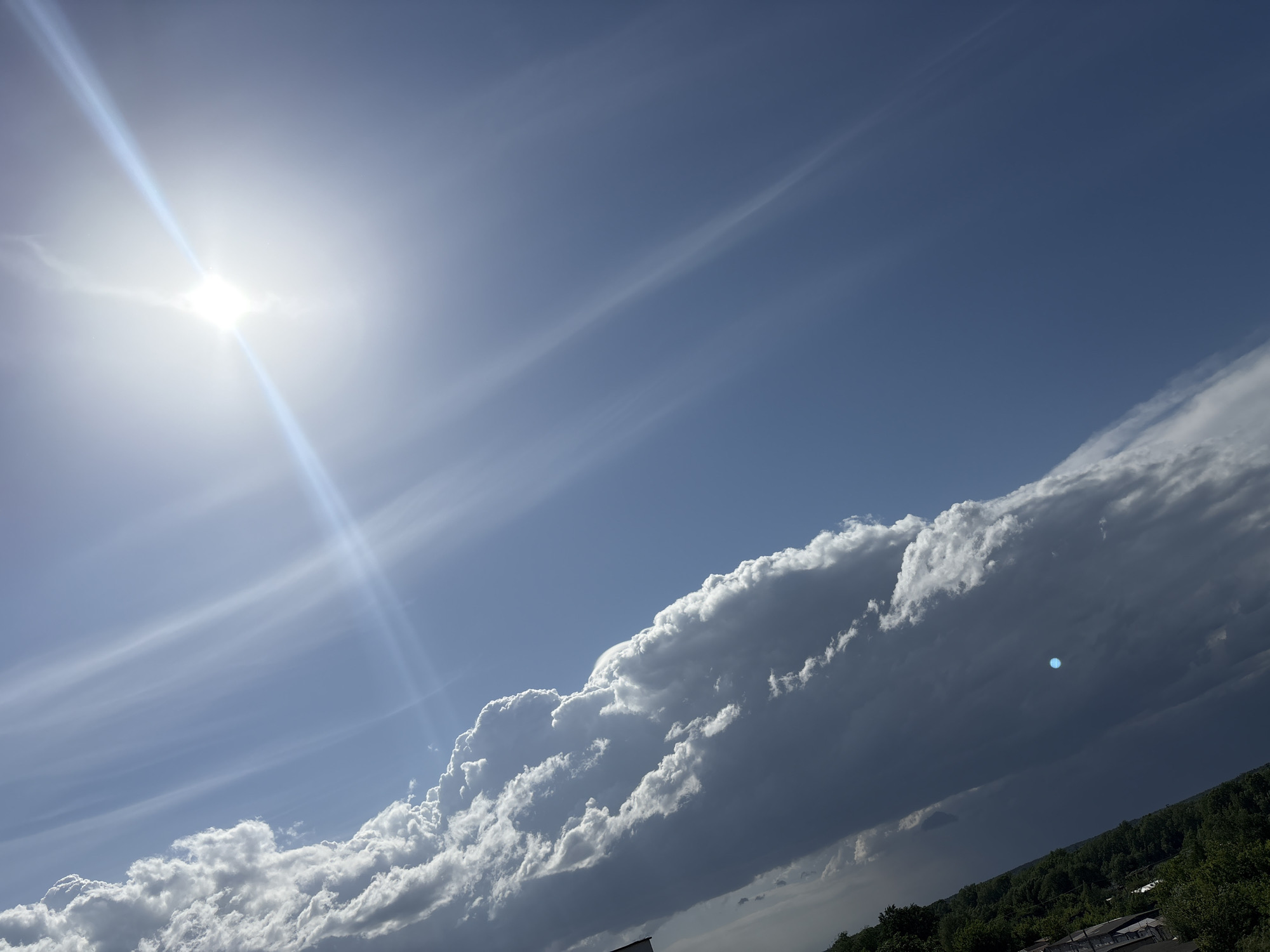 The image shows a bright, daytime sky over Chaikovsky, Russia. The sun is visible in the upper left, with lens flare extending downwards. Thin, wispy cirrus clouds stretch across the upper portion of the sky. Along the bottom right, a large cumulonimbus cloud mass dominates, transitioning from bright white at the top to dark gray below. The tops of trees and some buildings are visible beneath the clouds in the lower right corner.