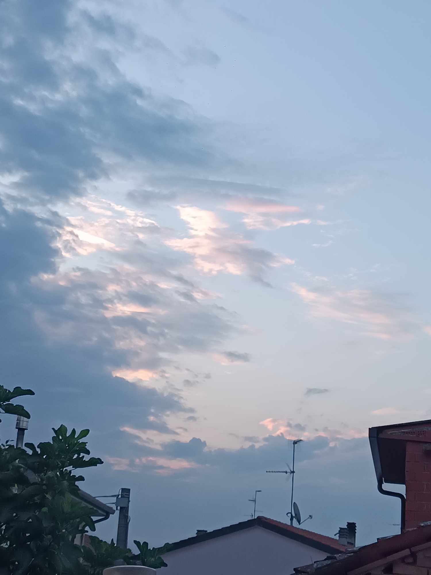 The image depicts a twilight sky over the city of Viadana, Italy. The sky is filled with a mix of dark blue and grey clouds, with streaks of pink and white where the last rays of sunlight are breaking through. In the foreground, the rooftops of several buildings are visible. To the left, the dark green leaves of a tree are prominent. To the right, a red brick chimney stands out against the sky. Various antennas and a satellite dish are mounted on the rooftops, suggesting a residential area. The overall scene conveys a calm and serene atmosphere of the transition from day to night.