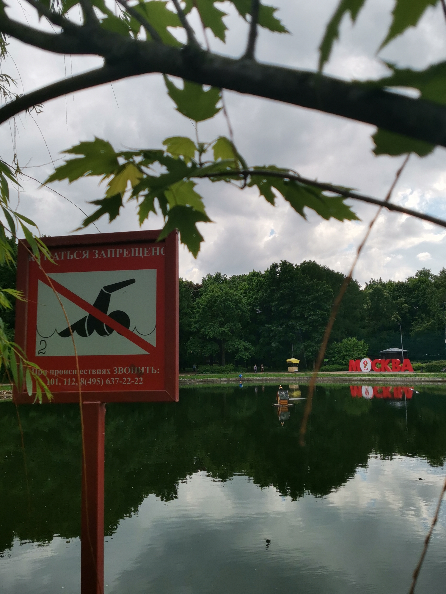 The image shows a calm lake with a "No Swimming" sign in the foreground, partly obscured by leaves. The sign is red with a white background displaying a swimmer crossed out with a red line. Below the image is text in Russian, along with telephone numbers. Across the lake, the red letters "МОСКВА" (Moscow) are visible, with a Russian flag design on the "O." A small wooden structure resembling a birdhouse is floating on the lake. The reflection of the sky and surrounding greenery are visible on the water's surface. The scene is taken during the day, with an overcast sky. The location is likely in a park in Moscow, Russia.