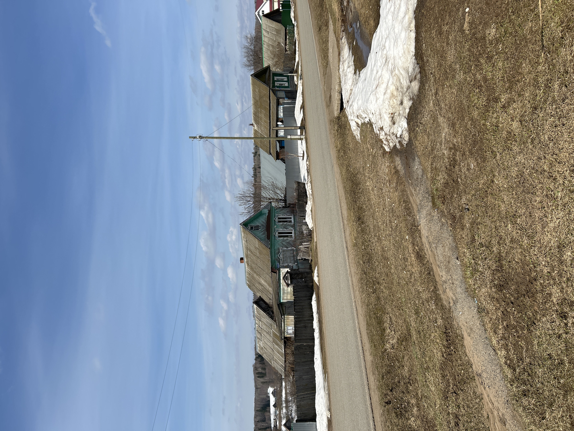 The image depicts a rural scene in Уральское, Russia. A narrow paved road stretches through the frame, bordered by dry grass and patches of melting snow. Several old, wooden houses line the road, showing signs of age and weathering. A wooden fence runs along the edge of one property. Power lines are visible in the sky, supported by a wooden pole. The sky is a clear blue with scattered clouds. The photograph suggests a quiet, possibly remote, village setting.