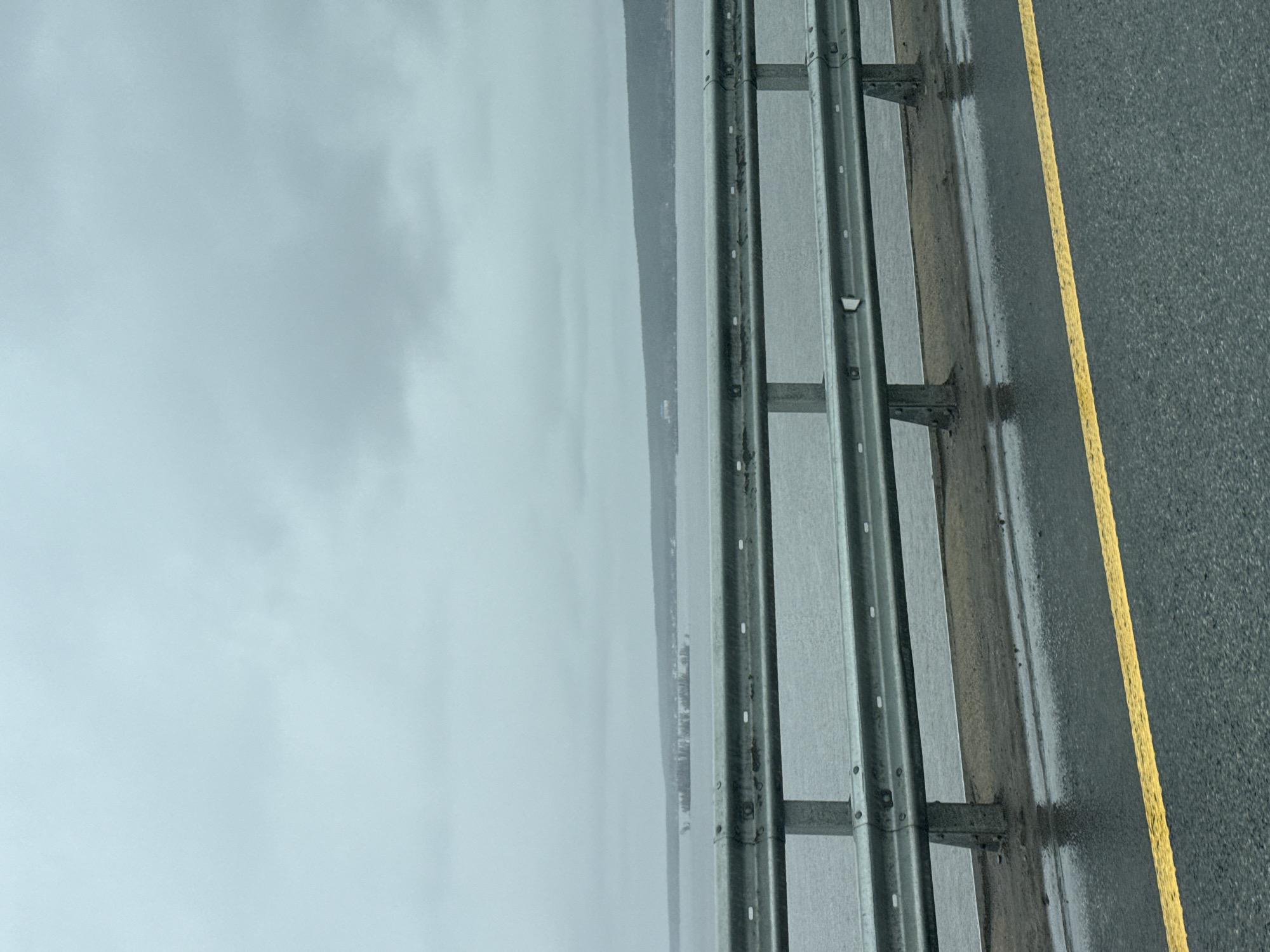 The image shows a view from a road on a foggy day. To the right of the frame is a grey asphalt road with a yellow line marking the edge. A metal guardrail runs along the edge of the road, separating it from the body of water. The body of water appears to be a lake or river, with a shoreline visible in the distance. The sky is overcast and grey. The location is presumed to be near a body of water in an urban or suburban area, based on the roadway and guardrail and a distant treeline and possibly buildings. There is no clear evidence to confirm that this image is located in Grebeni, Russia.