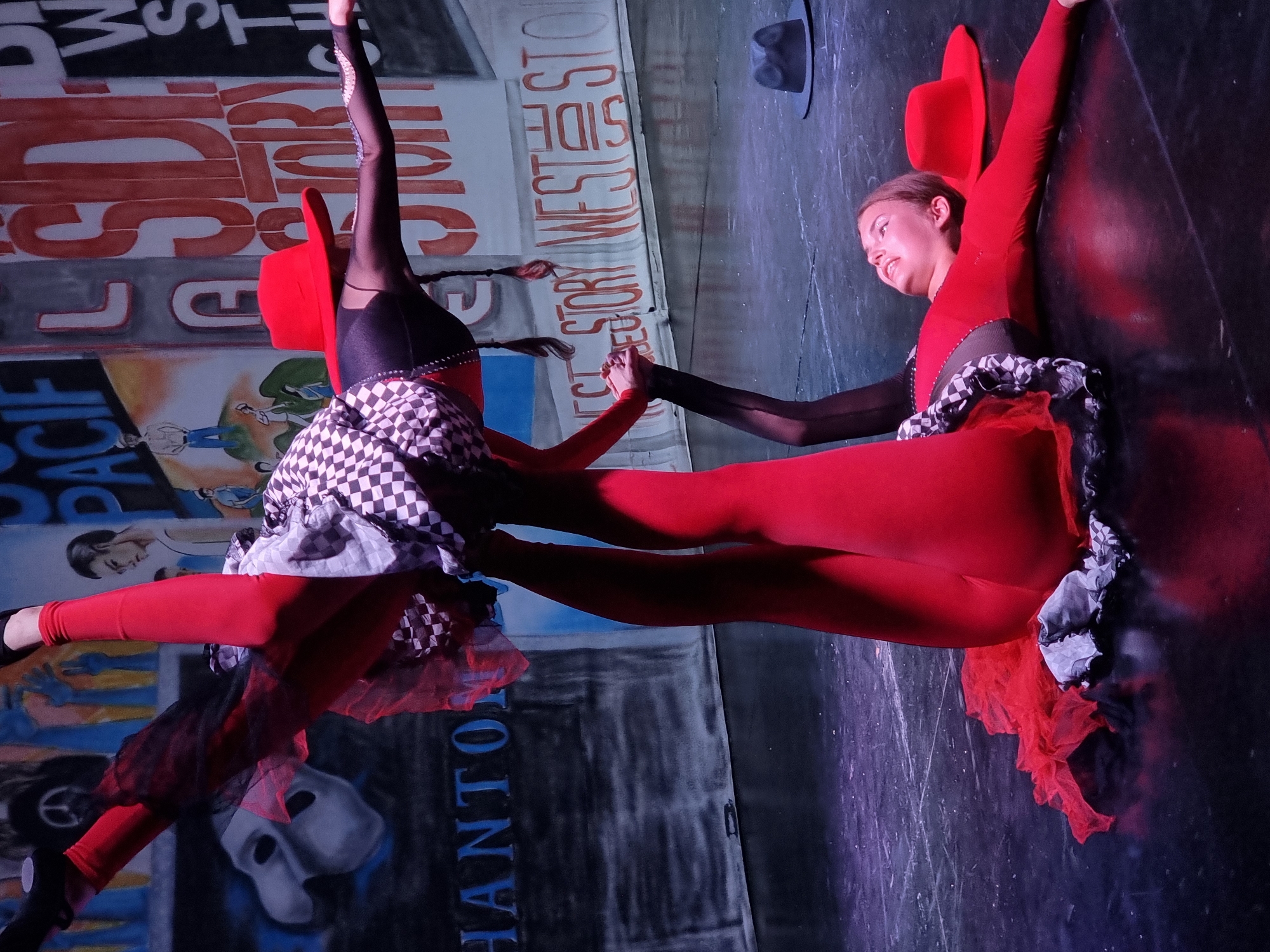 Two young women are performing an acrobatic dance routine on a stage. The woman on the bottom is lying on her back, wearing a red leotard and a red hat. The woman on top is balancing on the bottom woman's feet, wearing red leggings, a black and white checkered skirt, and a red hat. She is upside down, with her legs in the air. In the background, there is a painted backdrop with large letters that appear to spell out "WEST SIDE STORY". There is also a black fedora on the floor to the right of the frame. The stage is dark and appears to be made of wood. The lighting is dramatic, with spotlights highlighting the performers. The location is a stage in Manavgat, Turkey.