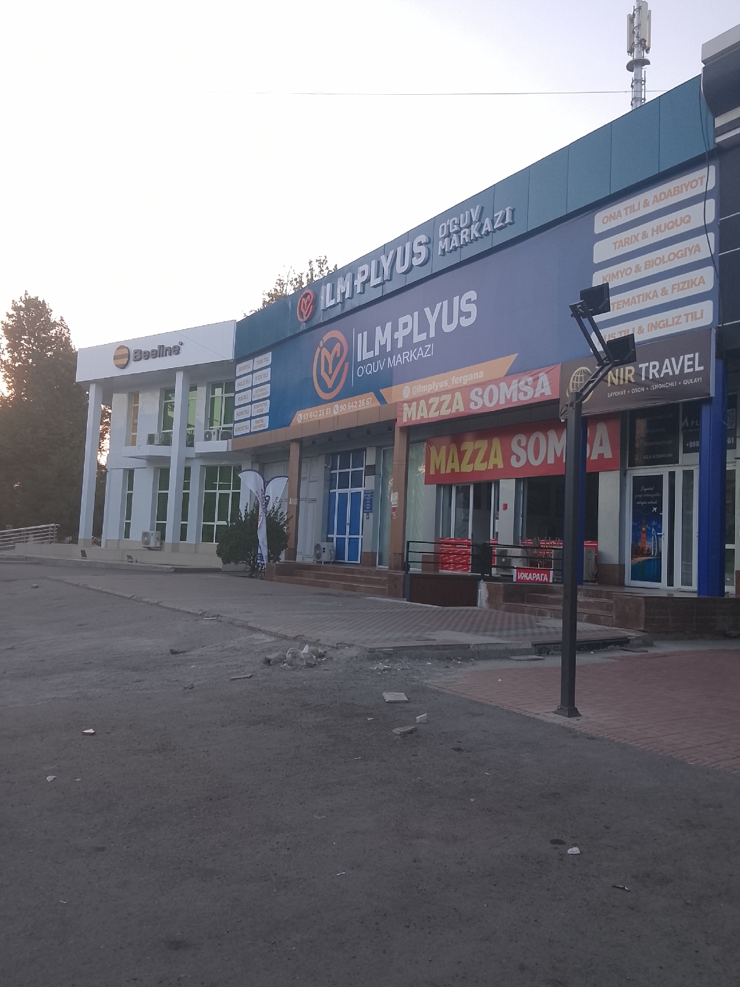 The image shows the exterior of a multi-story building with several businesses. The building is located on a street in Fergana City, Uzbekistan. On the left side of the building is a business with a "Beeline" logo. To the right of the Beeline store is a larger sign for "ILM PLYUS" with "O'QUV MARKAZI" written below it, suggesting an educational center. Below the "ILM PLYUS" sign, a banner advertises "MAZZA SOMSA." Further to the right, a sign for "NIR TRAVEL" is visible. The building has a modern design with large windows and visible air conditioning units. The street in front of the building is paved, and there are some debris on the ground. The sky above is clear and appears to be late afternoon or early morning.