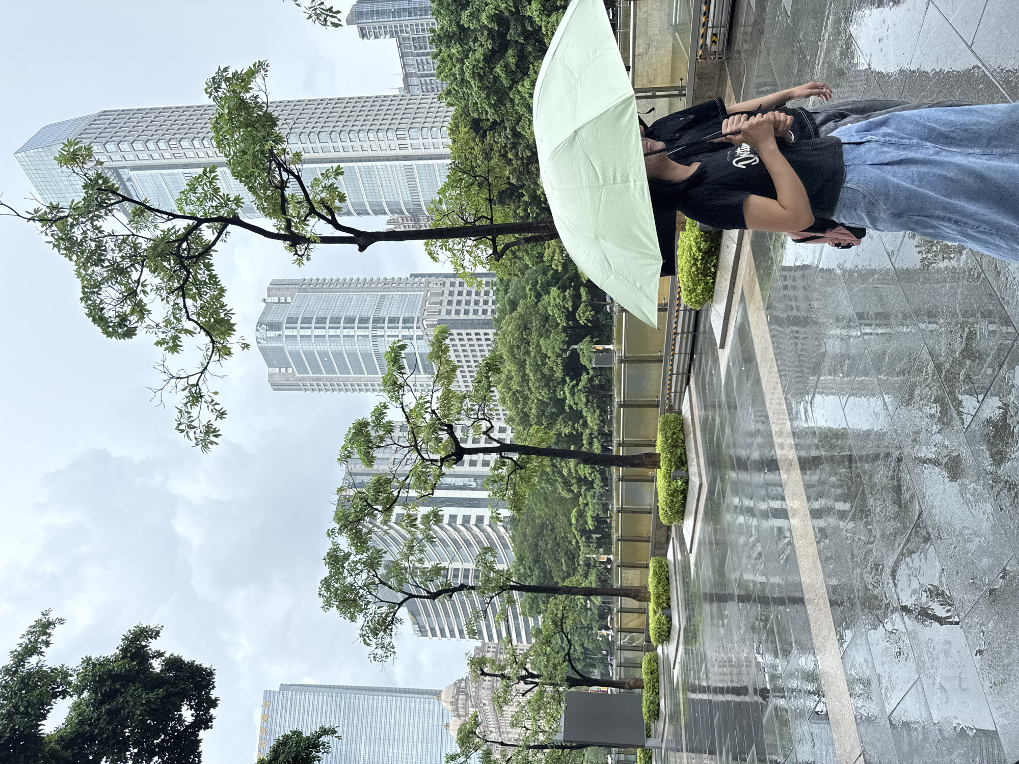 Two individuals are walking under a light green umbrella on a wet paved area in Tianhe District, China. The ground is reflective due to rain. In the background, several modern skyscrapers and lush green trees are visible. The sky is overcast. One person, a female, is wearing a black t-shirt and a light blue denim skirt, and has a pink backpack. The other person, a male, is wearing a black t-shirt and gray pants. They are walking away from the viewer.