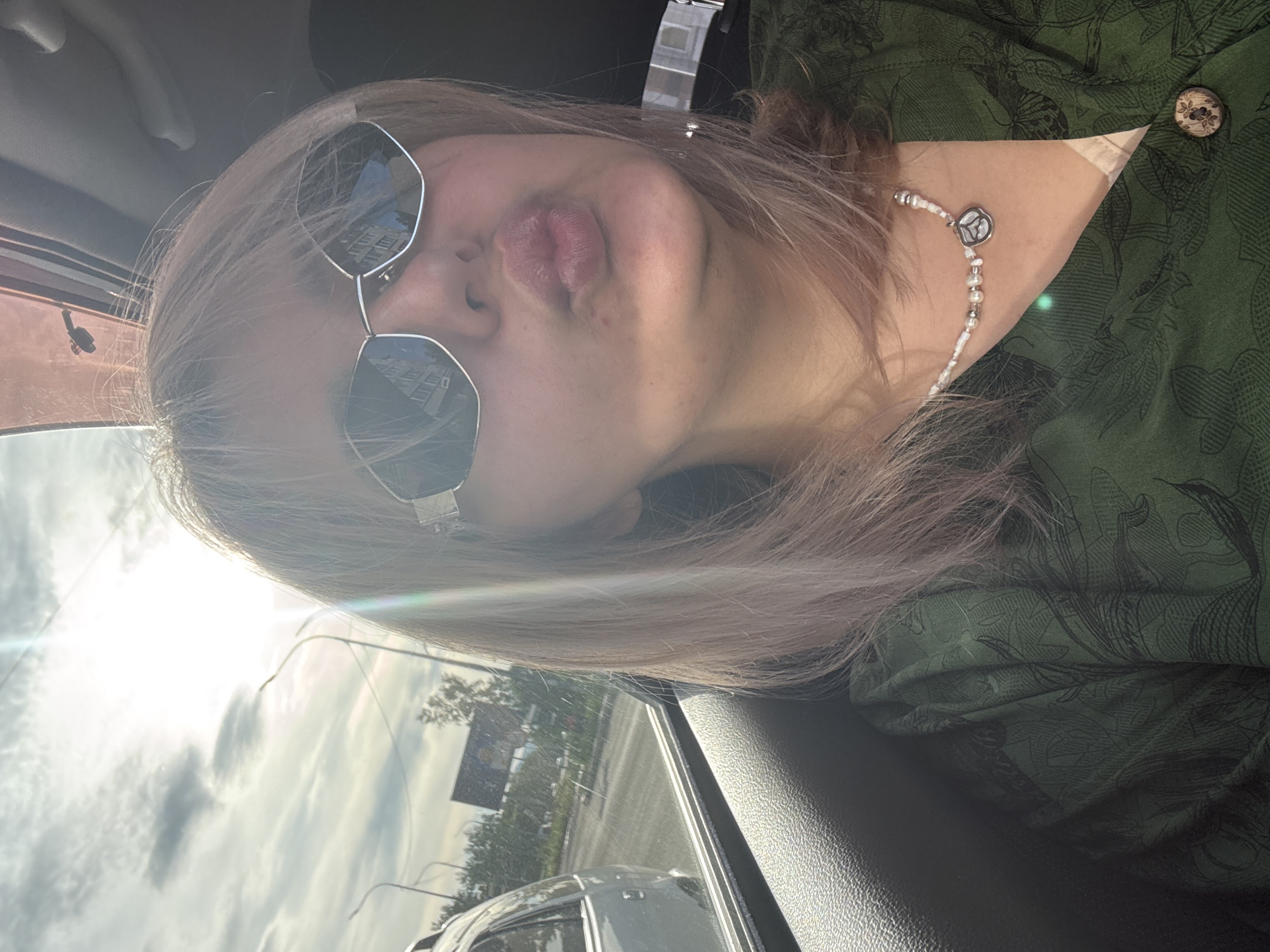 A blonde woman with a light skin tone is taking a selfie in a car, leaning forward to make a kissing face. She is wearing large, octagonal sunglasses with silver frames, a green patterned shirt, and a pearl necklace with a silver pendant. The sun is shining brightly through the car window, creating a lens flare. Outside the car, a street with trees and a billboard is visible. The interior of the car, including the door panel and ceiling, is also visible. The scene suggests the woman is traveling through the city of Birobidzhan, Russia.