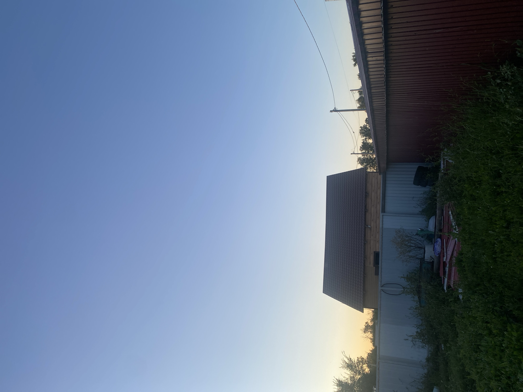 The image captures a clear, pale blue sky with a subtle gradient towards a lighter hue near the horizon, suggesting either dawn or dusk. Dominating the lower portion of the frame are various elements of a rural or suburban backyard. A wooden house with a dark, tiled roof is visible, partially obscured by a corrugated metal fence. This fence, painted in a dark reddish-brown, runs horizontally across the right side of the image. To the left of the house, a lighter-colored corrugated metal fence is present, with overgrown green grass and vegetation in the foreground. Among the debris near the lighter fence, a stack of black tires and some red corrugated metal sheets are discernible. In the background, a line of trees forms a silhouette against the sky. Utility poles and wires are also visible, stretching across the upper right quadrant of the image. The scene appears to be an outdoor area in Верхний Карбуш, Russia.