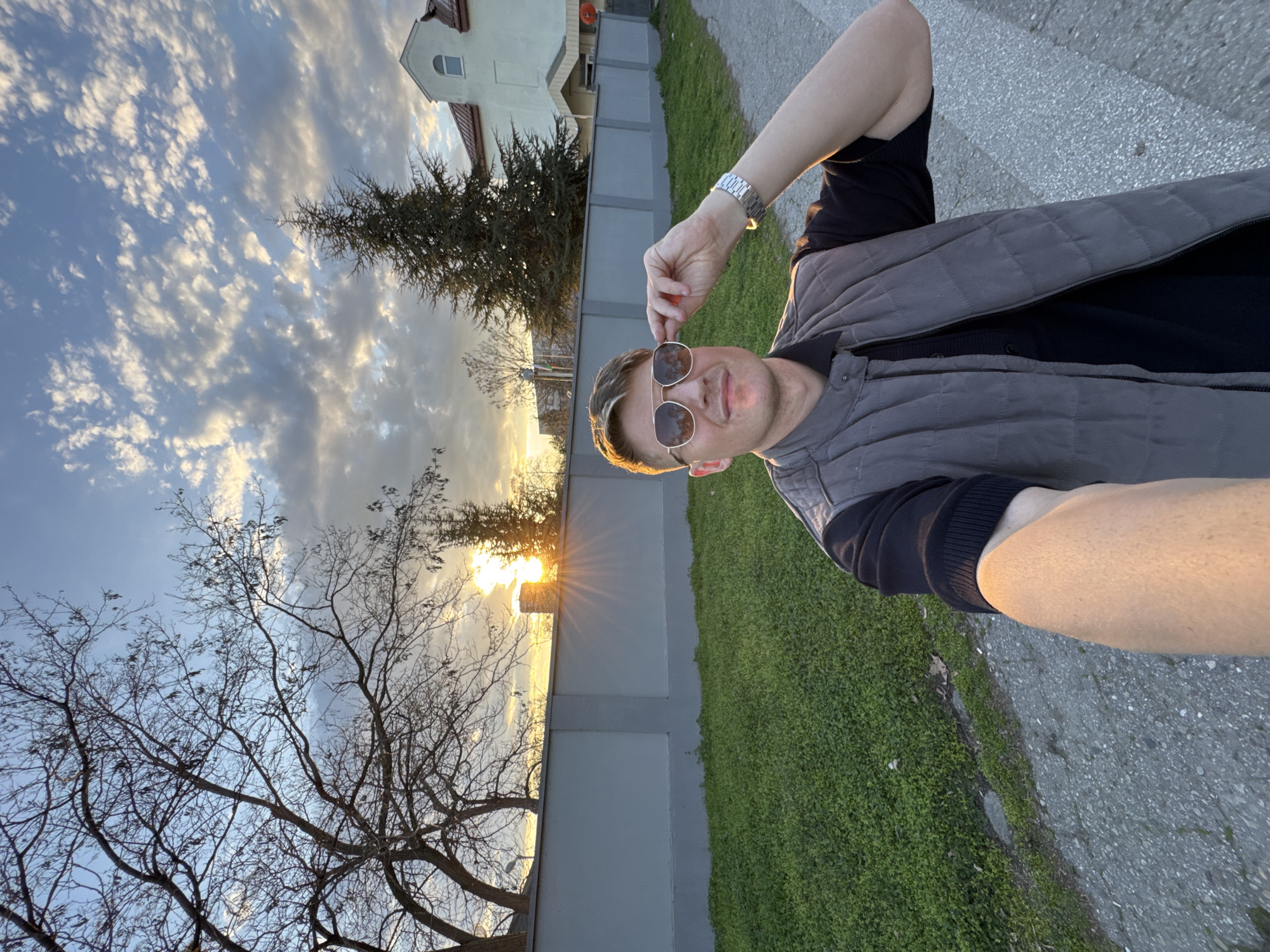 A man takes a selfie at sunset. He is wearing sunglasses, a black shirt, and a grey quilted vest. He is standing on a paved path next to a grassy area. A grey fence runs behind him, with trees and a building beyond. The sun is setting behind the fence, creating a bright glare. The sky is filled with clouds. I cannot confirm that this image was taken in Plovdiv, Bulgaria.