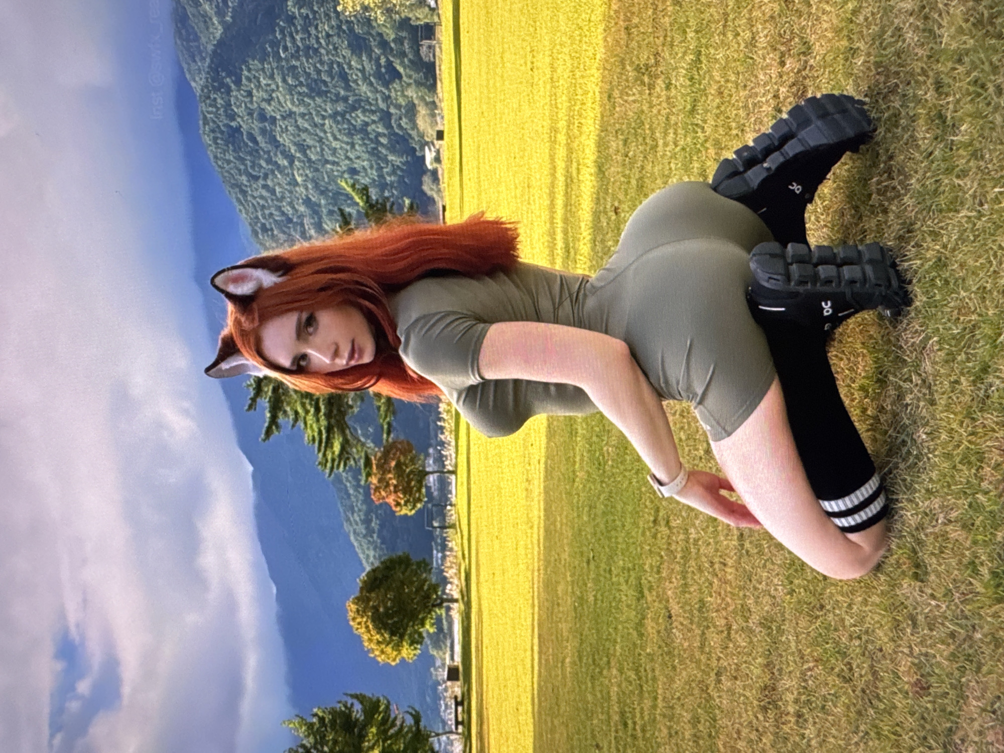 The image shows a person with long, reddish-orange hair and artificial fox ears, wearing a green outfit and black socks with white stripes. They are posing on a grassy field, with mountains and a body of water in the background. The sky is cloudy. The person is on their hands and knees, with their head turned to the side, looking at the camera. Their body is arched, and their legs are bent. The overall mood is playful and suggestive. There is text visible in the upper left corner of the image which reads "Insta: @swfk_rea".
