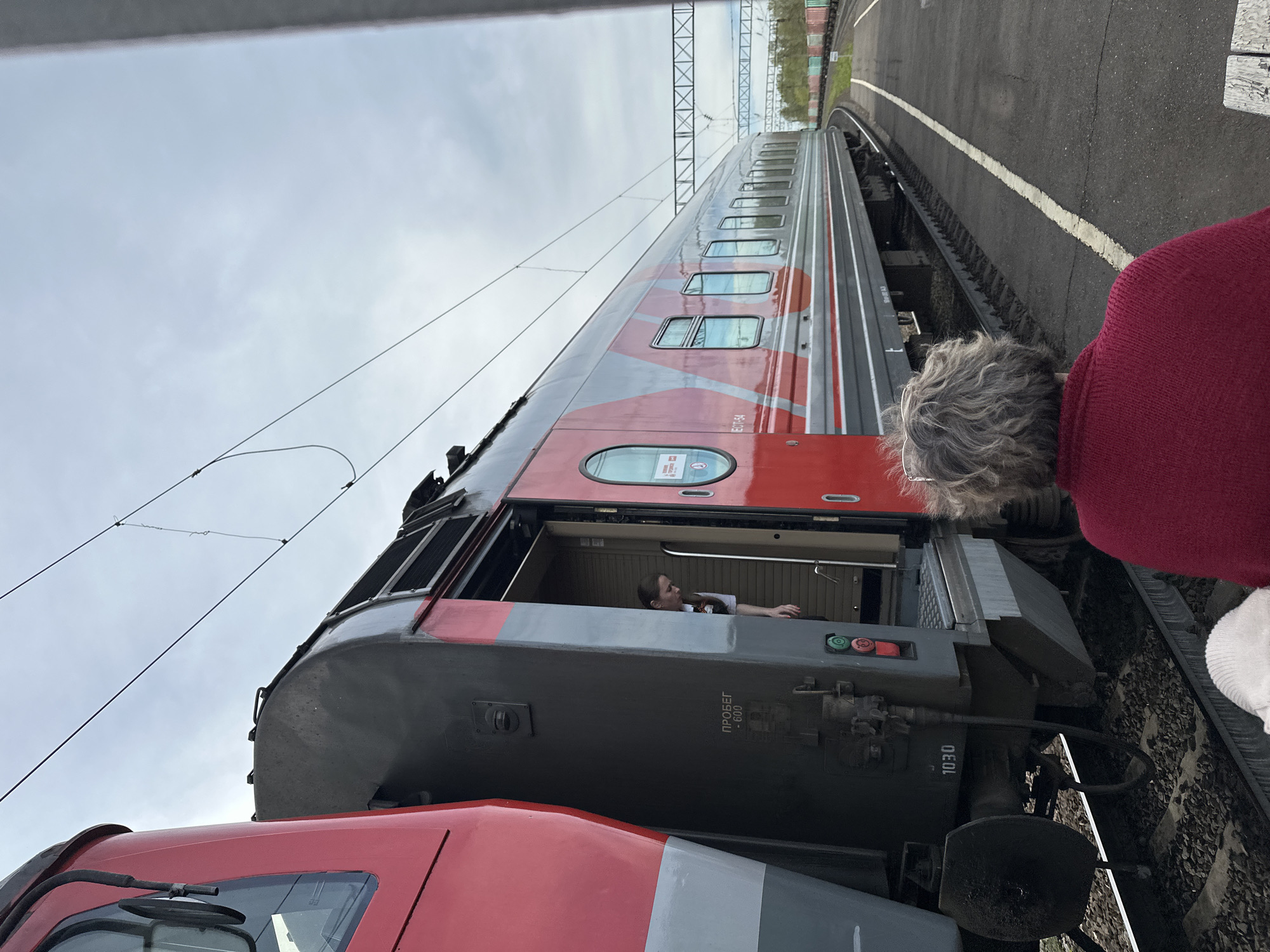 A red and grey train stands on tracks at a station in Кечёво, Russia. The train's door is open, revealing a woman inside the train car. A person with grey hair and wearing a red sweater is standing next to the train, looking toward the train and the woman inside. The sky is overcast, and the station platform is visible next to the tracks. Overhead power lines are also visible.
