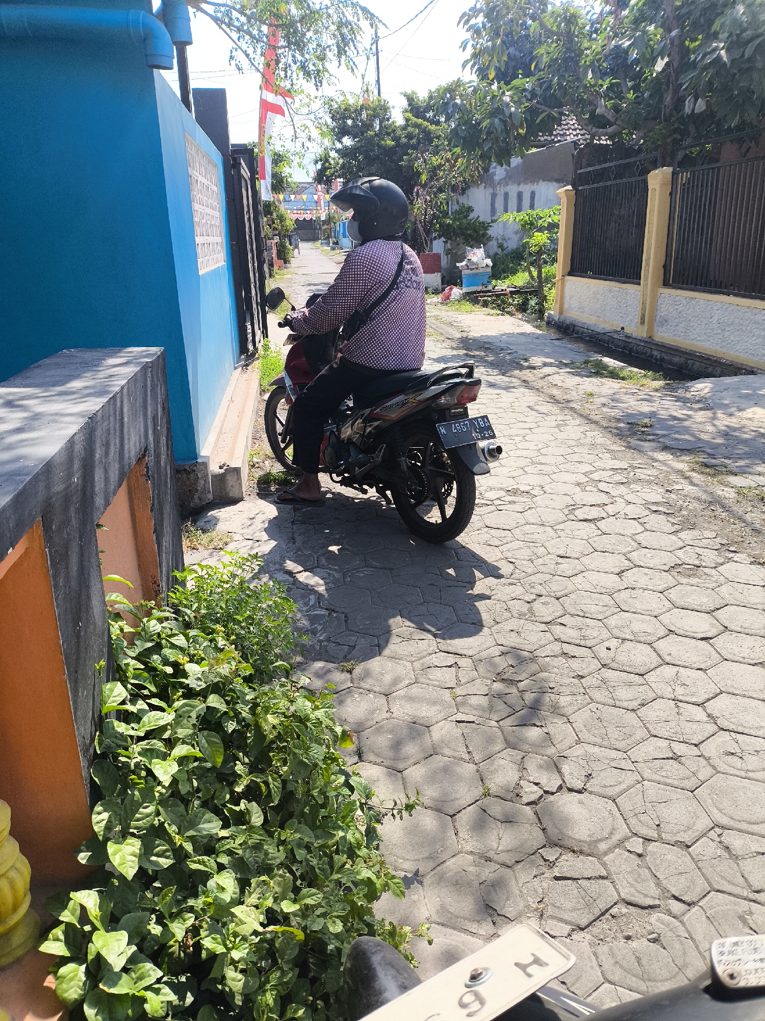 A person wearing a black helmet and a checkered long-sleeved shirt sits on a red and black motorcycle parked on a paved street in Lumajang, Indonesia. The street is lined with buildings and fences. To the left, a bright blue wall runs alongside the street, with a concrete structure in front of it. Lush green plants grow along the base of the blue wall. The motorcycle's license plate reads "N 4957 YBA." The ground is paved with hexagonal stones. Red and white Indonesian flags are visible in the background, suggesting a celebration or national event. The scene is illuminated by bright sunlight, casting shadows on the ground.