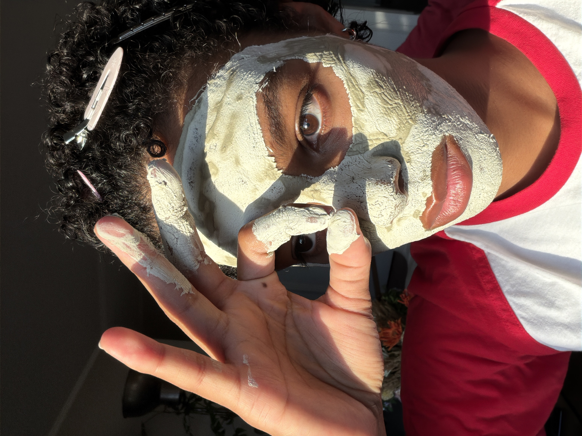 A close-up, low-angle shot shows a person with curly, dark hair, part of which is held back with pink hair clips. The person is wearing a clay mask, which is light green and cracked in places, covering most of their face, except for their eyes, lips, and nostrils. Their right eye is visible, brown, and looking towards the camera. Their lips are full and appear to be glossed. The person is wearing a red and white shirt. The light is coming from the left, casting shadows across their face and illuminating their left hand, which is raised and positioned in front of their face, with fingers covered in the clay mask. The background is dark and out of focus, with hints of greenery and possibly an interior space. The setting is not identifiable as Champs-sur-Marne, France.
