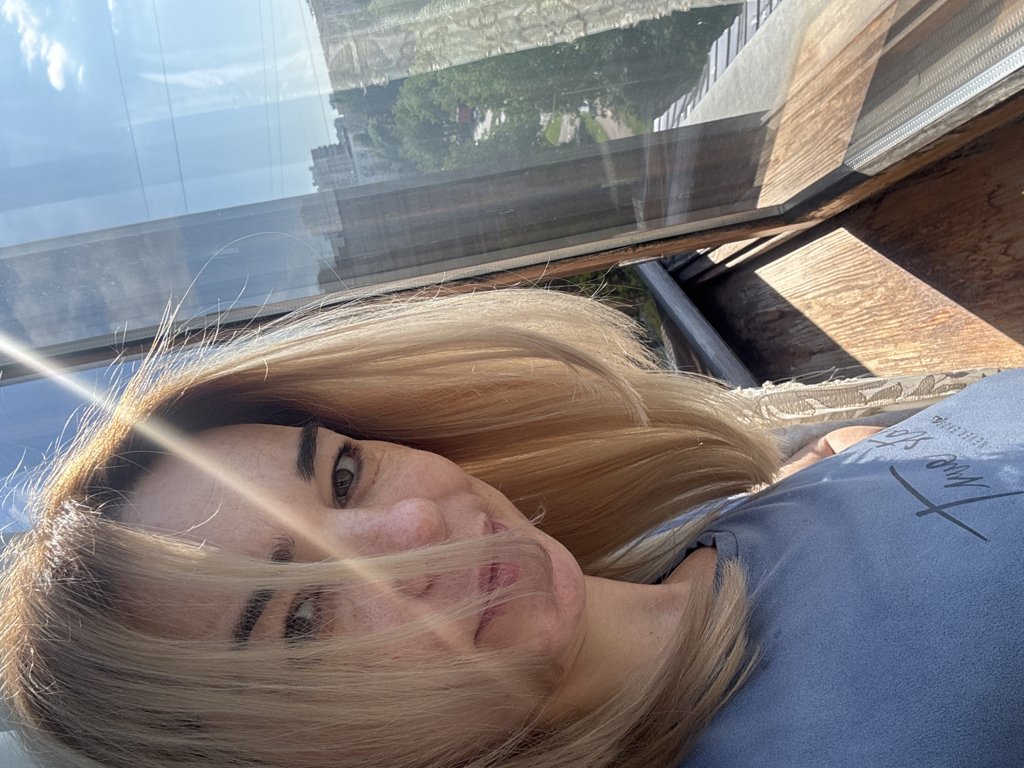 A woman with blonde hair is lying down, facing the camera. She is wearing a blue shirt with the text "NINE STONE" written on it. She is indoors next to a window. Outside the window there is a building with trees. The city is unknown.