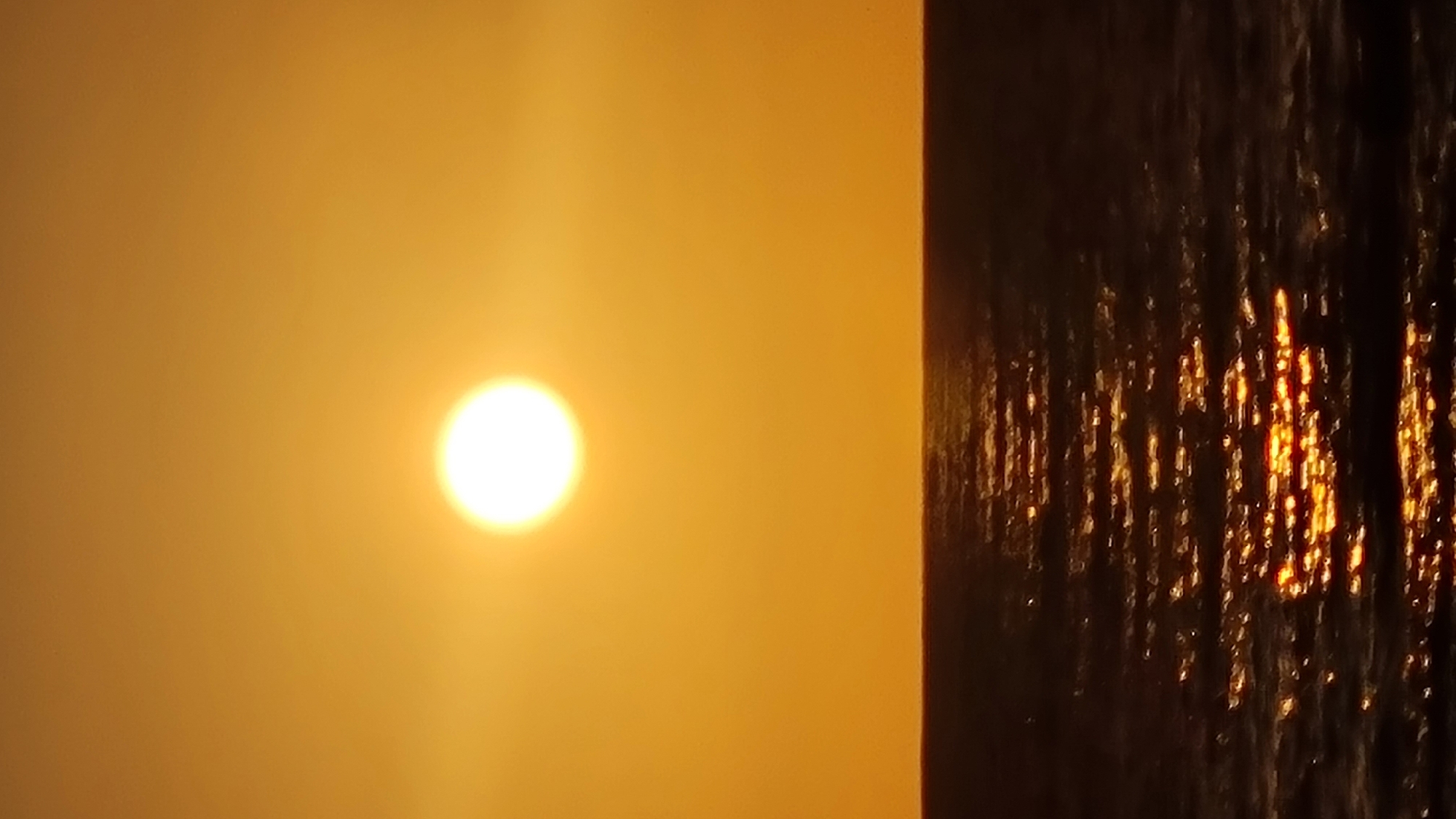 The image shows the sun, a bright circle, in a hazy, orange sky. To the right, there is a dark vertical strip with shimmering light reflections, likely a building or a waterfall reflecting the sunlight. The location in Nahariyya, Israel is not discernible from the image alone.