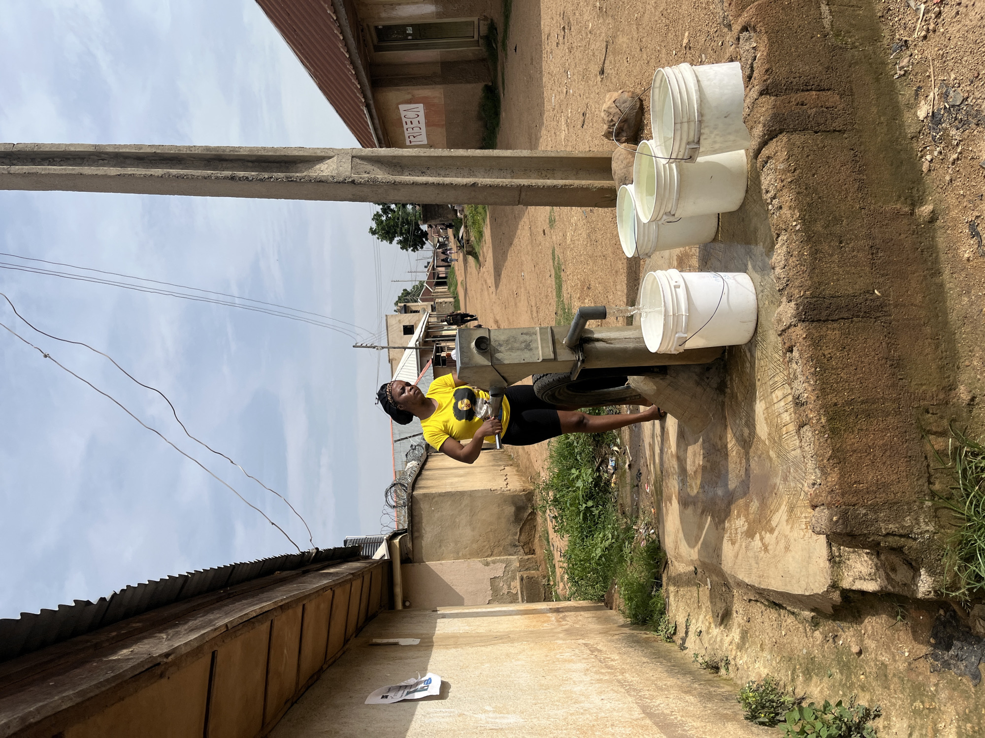 In Jos, Nigeria, a young woman stands by a manual water pump, operating it to fill buckets. She wears a bright yellow t-shirt and dark shorts, with a headscarf. Four white plastic buckets are positioned beneath the pump's spout, with water flowing into the closest one. The pump is made of concrete and metal. The environment is dusty and rural, with a dirt road leading into the distance. Buildings with corrugated metal roofs line the road. A concrete utility pole stands tall near the pump, and overhead electrical wires crisscross the sky. The scene suggests a daily task of water collection in a community.
