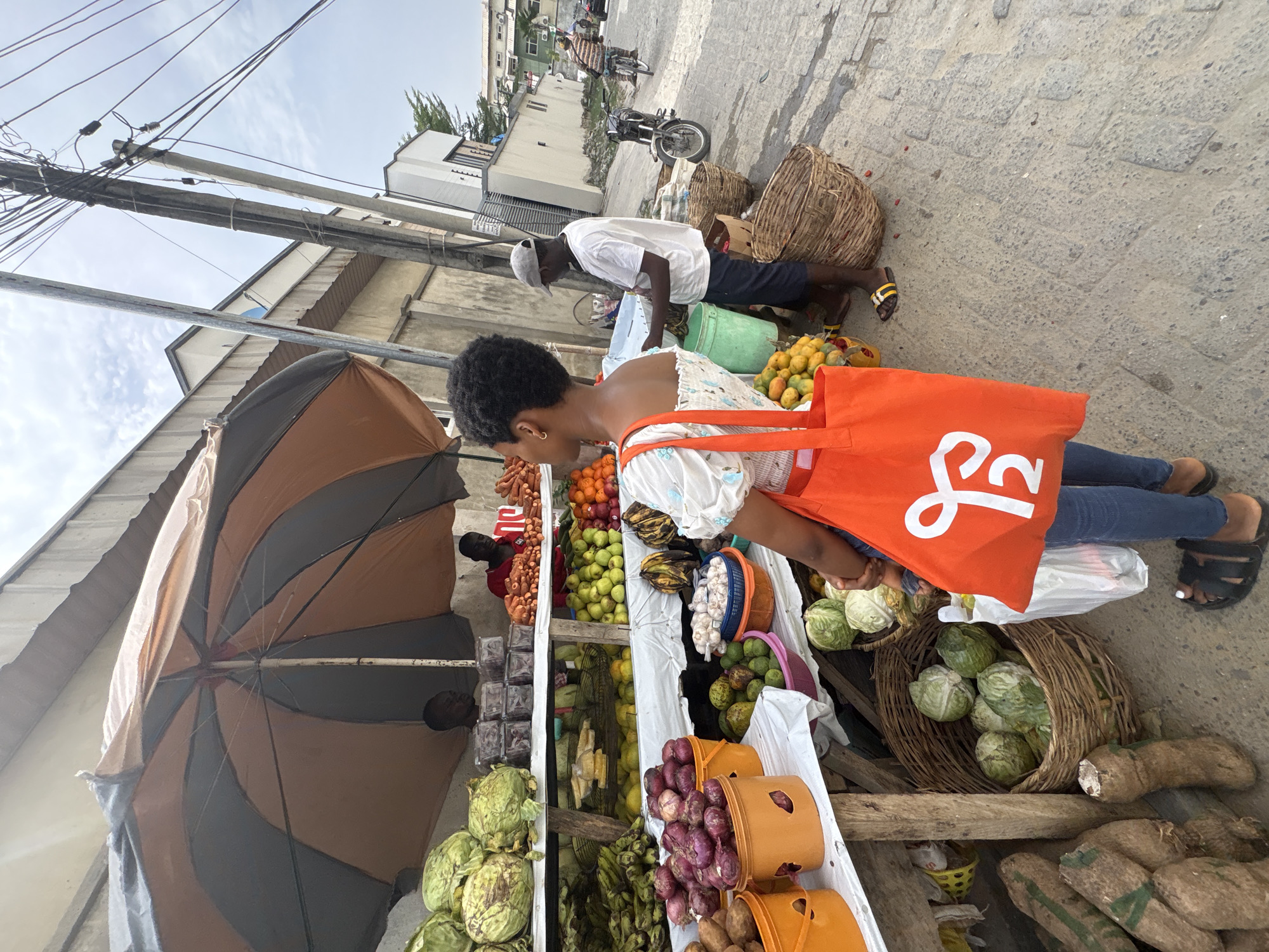 The image captures an active outdoor market scene on a paved street in Igboefon, Nigeria. In the immediate foreground, a female subject, observed from behind, wears a white off-shoulder top, blue jeans, and dark sandals. She possesses short, dark, curly hair and carries a bright orange tote bag with a white "P2" or "Y2" logo, along with a white plastic bag.
To the right, a market stall is set up under a large brown and dark gray striped umbrella. The stall features multiple wooden shelves draped with white fabric, laden with various fresh produce. Visible items include large green cabbages, red onions in orange plastic buckets, carrots, oranges, green apples, bananas, mangoes, avocados, limes, and garlic. Yams are stacked on the ground beside the stall. Two male vendors are present at the stall; one on the right wears a white t-shirt, dark shorts, and a light-colored cap, appearing to attend to goods, while another, partially obscured, wears a red shirt deeper within the stall. A third male vendor is faintly visible on the left side of the stall, behind produce.
The street surface consists of rough, gray concrete pavers. A silver motorcycle is parked on the right side of the street beyond the market stall. Further down the street, a male on a bicycle, wearing a striped shirt, rides away from the camera. Multiple utility poles with a dense network of black electrical or communication wires extend into an overcast sky. Buildings with light-colored walls and security fencing are visible along the street in the background. Large woven baskets rest on the ground near the market stall.