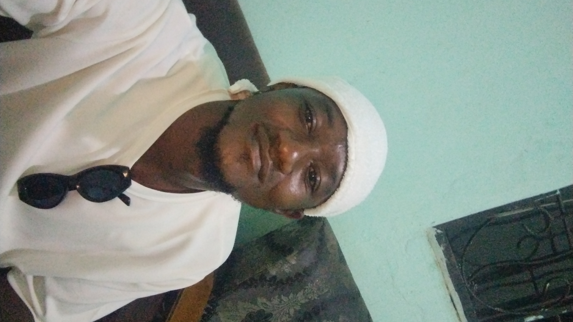 A man wearing a white beanie and a white t-shirt is pictured in a selfie. He has dark skin and a beard and is wearing sunglasses clipped to his shirt. The background appears to be a room with a light blue wall and a window with a dark metal grate. The overall impression is of an indoor setting, likely a residential or personal space. There are no clear indicators that this specific location is in Jos, Nigeria.