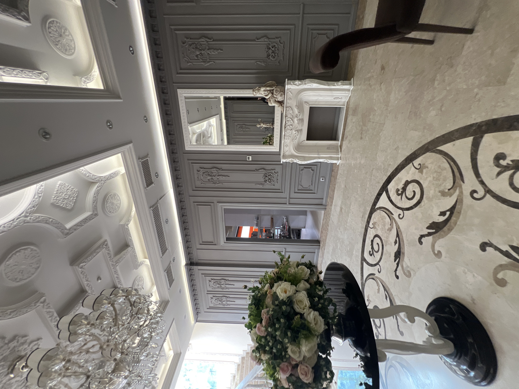 This image captures a luxurious and ornate interior space, located in Yerevan, Armenia, characterized by its classical design elements and elegant furnishings. The scene, viewed as if upright, showcases a grand room, possibly a hallway, dining area, or reception space.
The ceiling is a focal point, featuring elaborate white plasterwork with intricate moldings, recessed spotlights, and linear LED strip lighting that illuminates the details. A large, multi-tiered crystal chandelier hangs prominently, adding to the room's opulence. The walls are adorned with light grey paneling, rich with classical-style moldings and decorative friezes.
Towards the background, a majestic white fireplace mantel, highly sculpted with classical motifs, stands beneath a large, ornate mirror. A cherubic or putto statue rests on the mantelpiece. The mirror reflects parts of the ceiling and an opening into what appears to be a modern kitchen or serving area with stainless steel appliances and orange accents, suggesting a functional space beyond the aesthetic grandeur.
In the foreground, a lush floral arrangement of white and light pink roses with green foliage sits on a dark, glossy table with a sculptural white and black base. Partially visible to the right, a dark wooden chair with curved armrests is positioned on a light-colored marble or polished stone floor. The floor itself is accented with an intricate dark grey or black inlay pattern, forming elegant scrollwork. Natural daylight streams in from a large window on the left, revealing a glimpse of greenery and a staircase outside.
No people are visible, and no specific activity is taking place, creating a serene and ready atmosphere. The overall impression is one of sophistication, grandeur, and meticulous attention to classical detail, typical of a high-end establishment or a luxurious private residence. There is no visible text in the image.