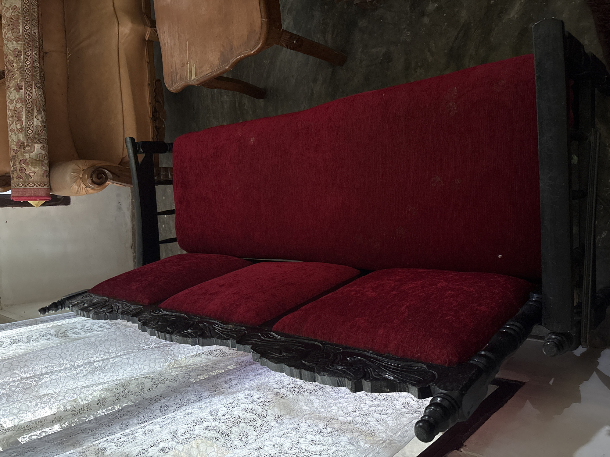 A dark wooden, intricately carved couch with plush red upholstery is positioned to the left of the frame. The couch has three visible seat cushions and appears to be part of a larger set. To the right of the couch is a wooden coffee table with a light brown, worn surface and carved legs. In the background, a tan leather sofa is visible, topped with a rolled-up rug that has a patterned border. A white lace curtain hangs on the left side of the image, covering a window. The floor appears to be concrete. The scene is indoors, in what seems to be a living room or parlor. The location is identified as Beunyot, Indonesia.