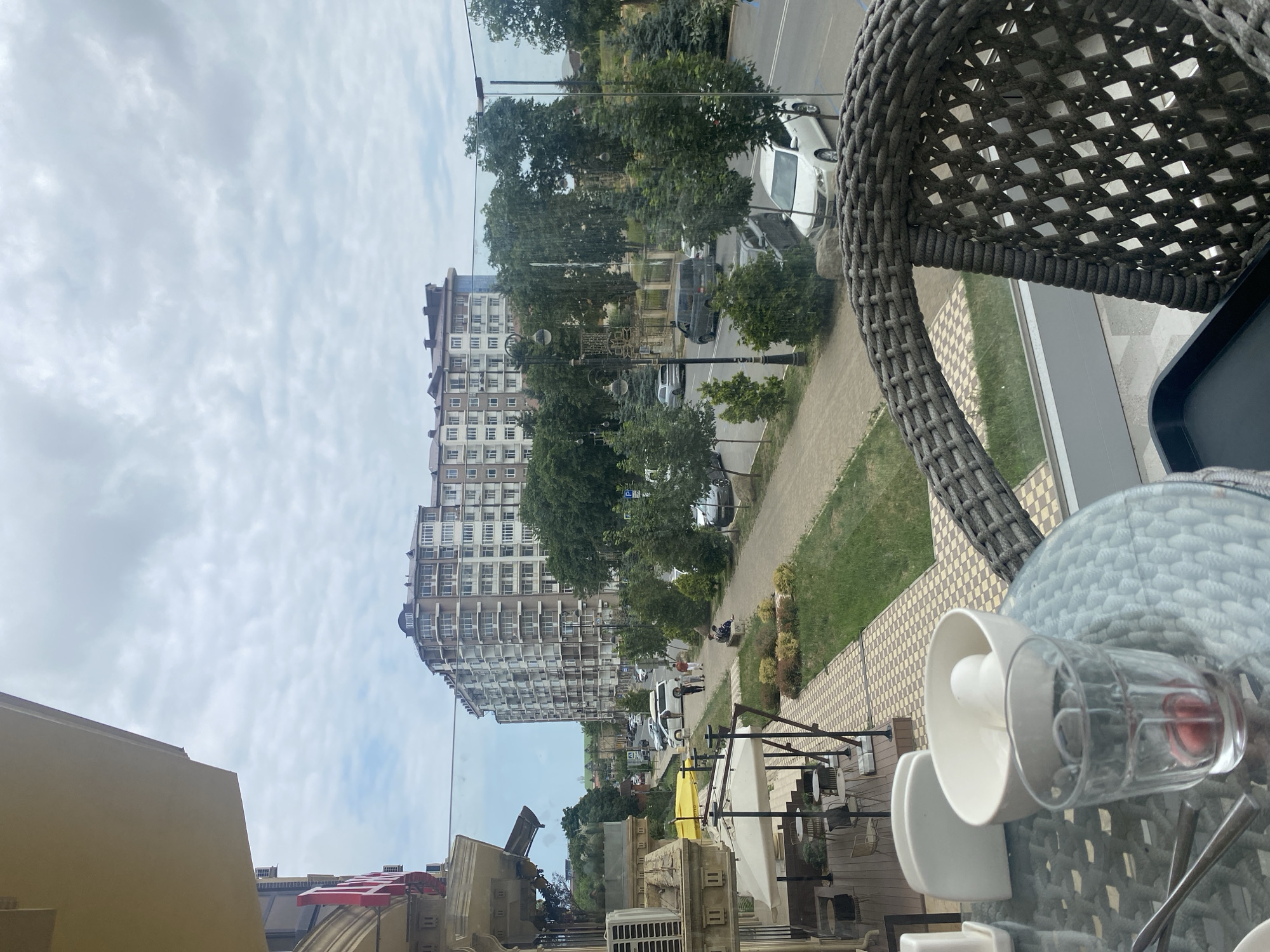 The image captures a view from a cafe or restaurant in Derbent, Russia, overlooking a street and a large, modern apartment building. In the foreground, a table with a glass, a white bowl containing two white objects, and a napkin holder is visible. A woven chair is positioned next to the table. Through a glass partition, a sidewalk with a checkered pattern leads to a green grassy area. Several trees are planted along the sidewalk and road. A street with parked cars and a few moving vehicles is depicted, including a white sedan and a dark van. A few people are walking on the sidewalk, and one person is seated on a bench. The sky is overcast with clouds. The architecture of the apartment building suggests it is a contemporary structure with multiple balconies. The overall scene portrays a common urban environment in Derbent during daylight.