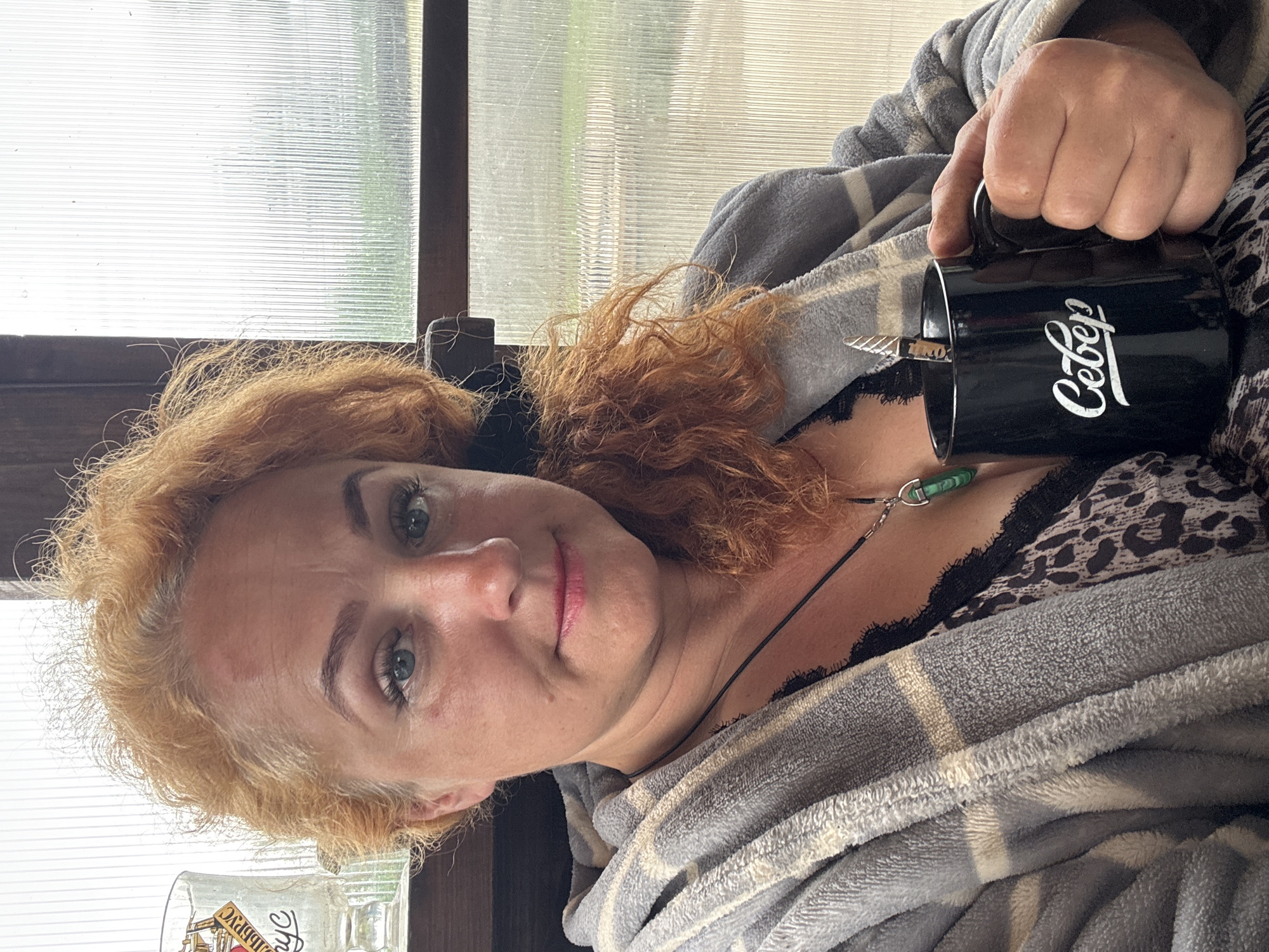 In a close-up, selfie-style shot, a woman with curly auburn hair and blue eyes sits indoors, holding a black mug in her right hand. She is wearing a grey and white plaid robe over a black leopard-print top with lace trim. A pendant necklace with a green stone hangs around her neck. The background consists of a ribbed translucent window with a view of greenery outside, suggesting a rural or semi-rural setting, possibly within a greenhouse or enclosed porch. To the left of the woman, partially visible, is a clear glass with text that appears to be in Cyrillic. The lighting is soft and diffused, indicating an overcast day or indoor illumination. The overall impression is one of a casual, relaxed moment. The text on the mug reads "Север", which translates to "North" in Russian. The woman's location is stated to be in the city of Chaikovsky, Russia.